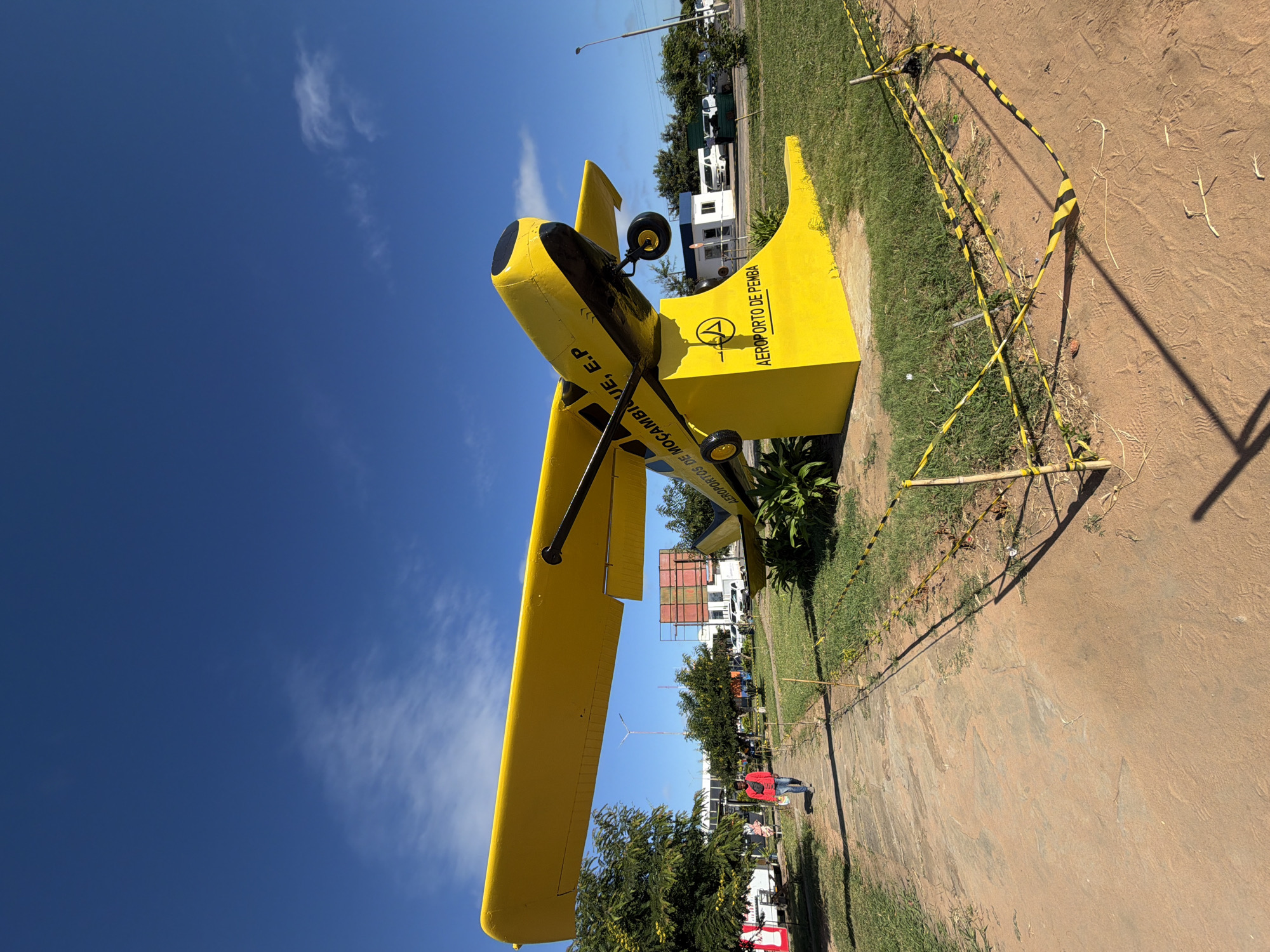 The image, oriented correctly, displays a bright yellow biplane-style aircraft on static display outdoors under a clear blue sky with wispy white clouds. The aircraft is predominantly yellow with black accents on its engine cowling, propeller, and some structural elements. On its fuselage, partial text reads "...DE MOÇAMBIQUE, E.P.", while the tail clearly shows "AEROPORTO DE PEMBA" along with a circular logo containing the letter 'A'. This identifies the location as Pemba Airport in Mozambique.
The aircraft is situated on a patch of green grass, next to a reddish-brown dirt path. A barrier made of yellow and black striped tape, supported by thin poles, surrounds the display. Long shadows are cast across the dirt, suggesting the photograph was taken during either the morning or late afternoon. In the background, beyond the aircraft, are various trees, a small white building, and other structures, including a white van and a dark green container. One person, dressed in a red shirt and dark trousers, is visible walking in the mid-ground near the plane's tail. The scene appears sunny and clear.