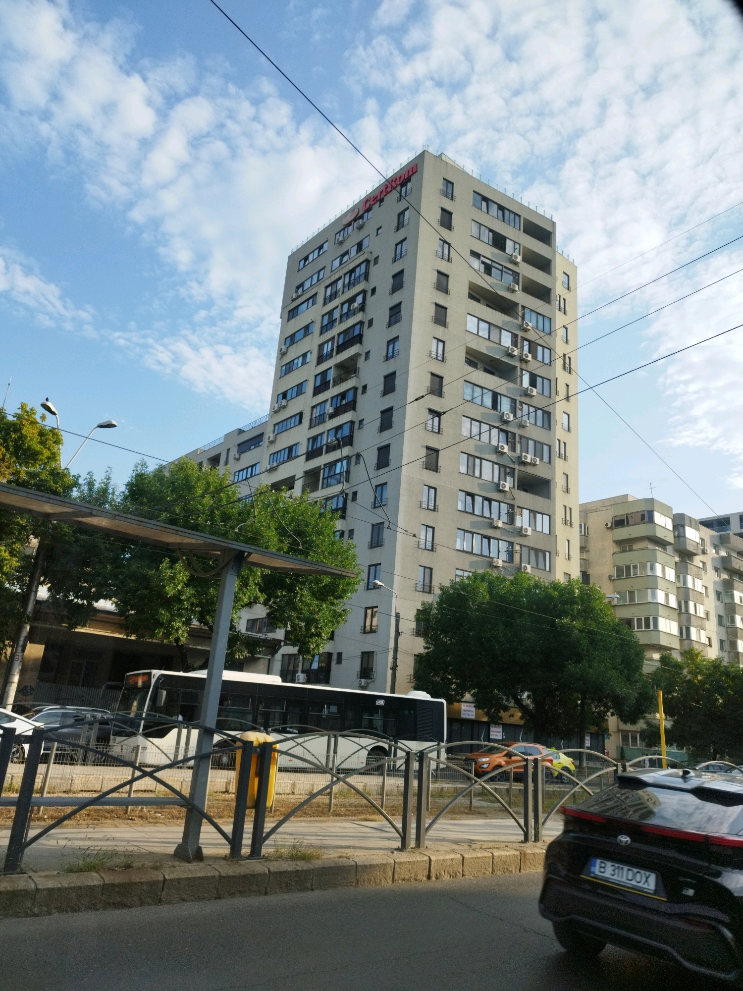 The image captures a street view in Bucharest, Romania, under a partially cloudy sky. In the foreground, a black car with license plate B 311 DOX is visible, partially obscuring a street with a sidewalk and a row of metal barriers. Behind the barriers, a bus and other vehicles are present. Further back, a tall, light-colored apartment building dominates the scene, with its upper facade featuring the red logo "Certikon". Adjacent to this building is another, shorter apartment building, also appearing to be residential. Lush green trees are interspersed between the buildings and along the street. The overall impression is of a typical urban environment in Bucharest.