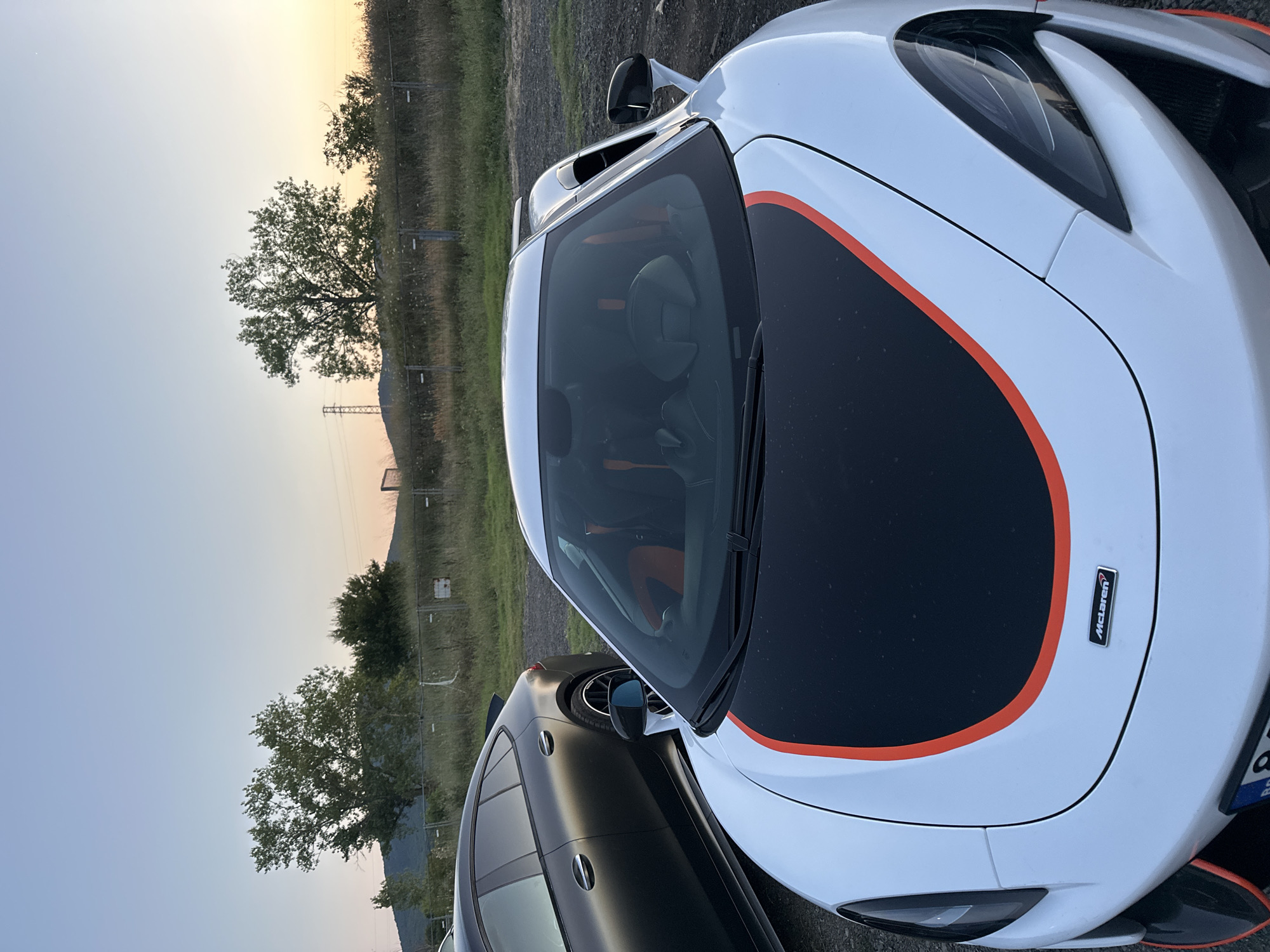 The image shows a close-up of the front of a white McLaren sports car, with a black hood featuring an orange stripe. To the left of the McLaren, a dark-colored sedan is parked. The vehicles are on a gravel surface, with dry grass and a fence in the background. Trees and a distant transmission tower are visible against a pale, sunset sky. The scene is located in Sozopol, Bulgaria.