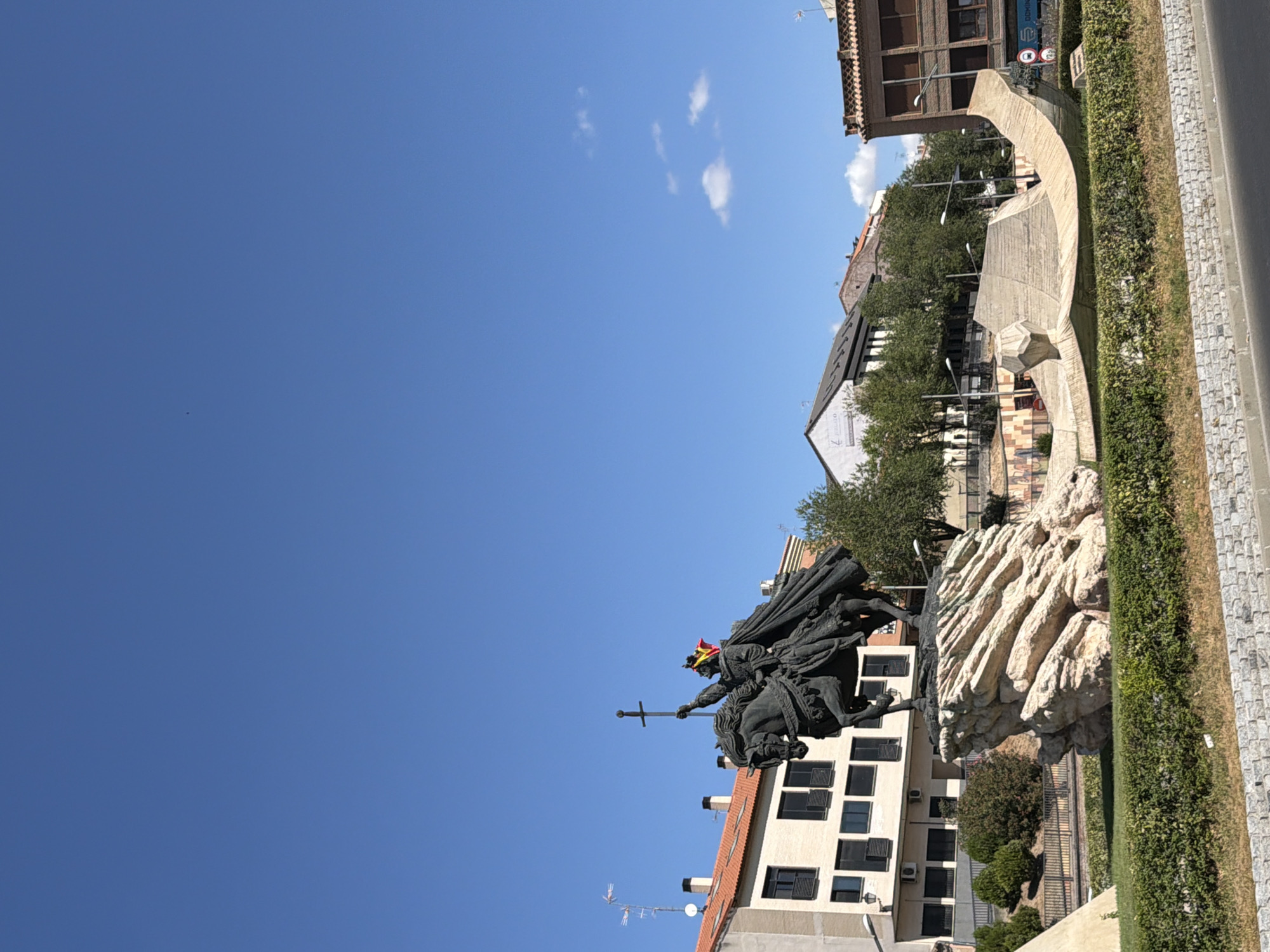 The image shows an outdoor monument featuring a bronze equestrian statue of a king, identified as King Alfonso VI of León and Castile, holding a sword aloft. The statue is placed on a rocky base. Behind the statue, several buildings are visible, including a light-colored building with dark windows and a red-tiled roof, and a darker brick building to the right. Trees and streetlights are also present. The scene is set against a clear blue sky with a few small clouds. The monument is located in Toledo, Spain.