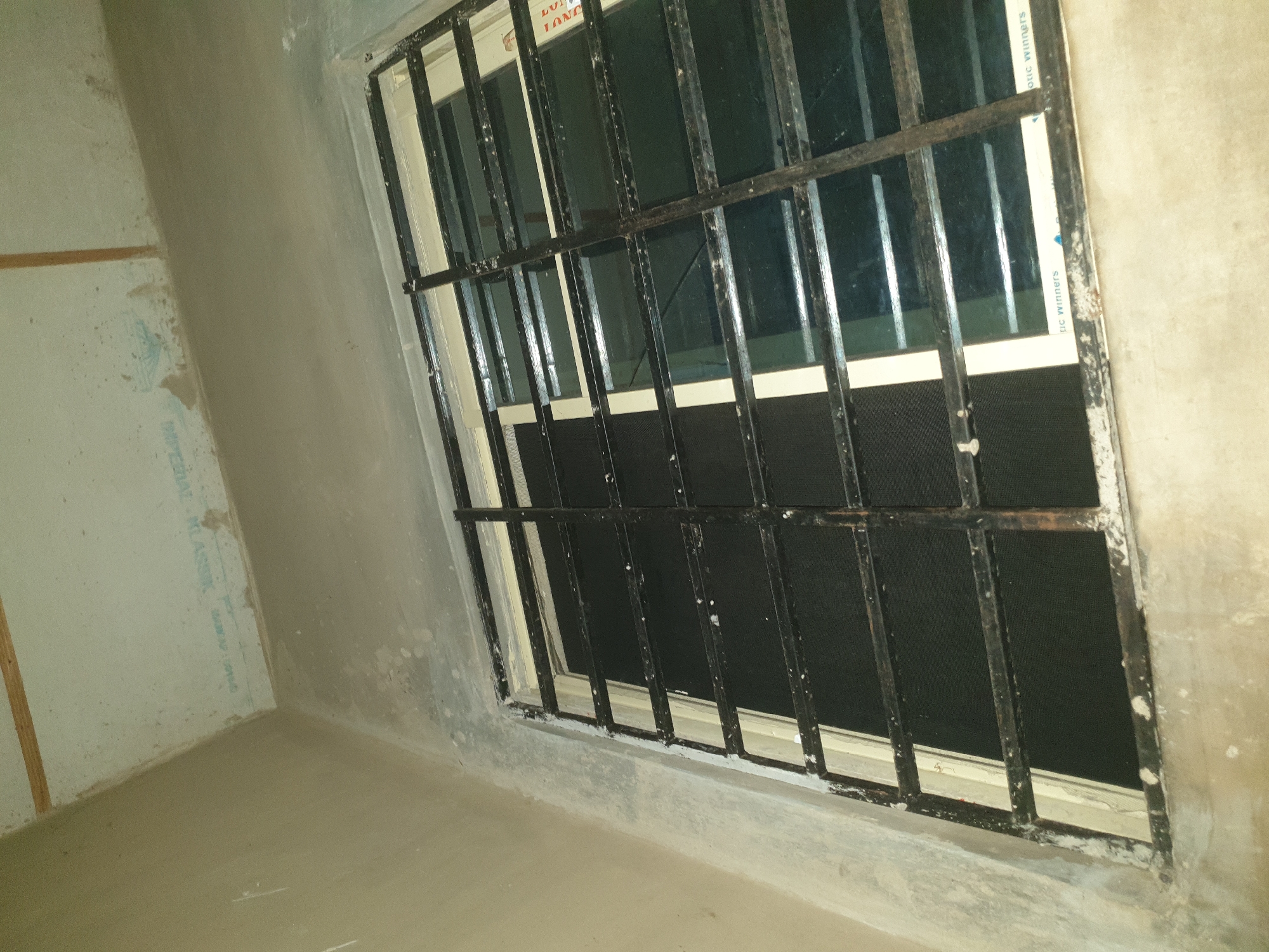 The image shows a view of a window with metal bars, seen from a low angle looking upwards. The window is set into a concrete wall. The window itself appears to be double-paned and has black mesh screens behind the bars. There is some white trim around the window frame. The ceiling is visible above the window and appears to be unfinished or under construction, with exposed wooden beams and rough concrete. The overall environment suggests an interior space that is either unfinished or in a state of disrepair. There are no people or animals visible in the image. The location is described as Unknown, Unknown.