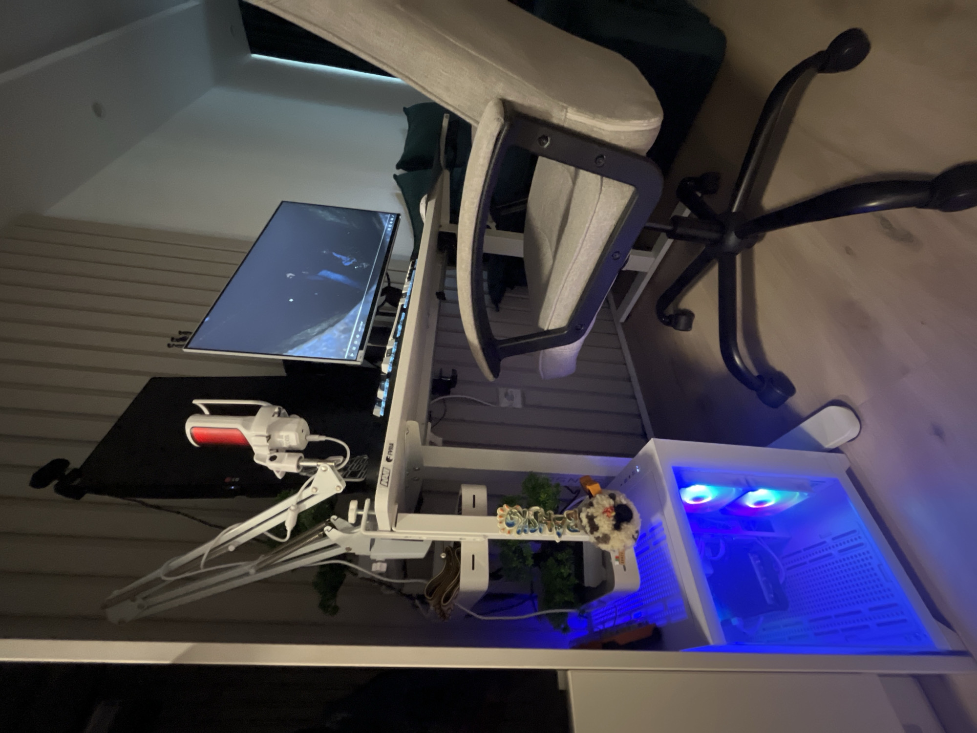 The image displays a modern computer setup in what appears to be a living space, possibly in Varna, Bulgaria. The primary focus is a white adjustable standing desk with a computer monitor, a microphone on a boom arm, and a keyboard. Below the desk, a white computer tower with blue LED lighting is visible. Adjacent to the desk is a light-colored office chair with a black base and wheels. The background features a wall with vertical paneling and a light-colored ceiling. A dark green sofa is partially visible to the right. The overall impression is of a functional and contemporary workspace.