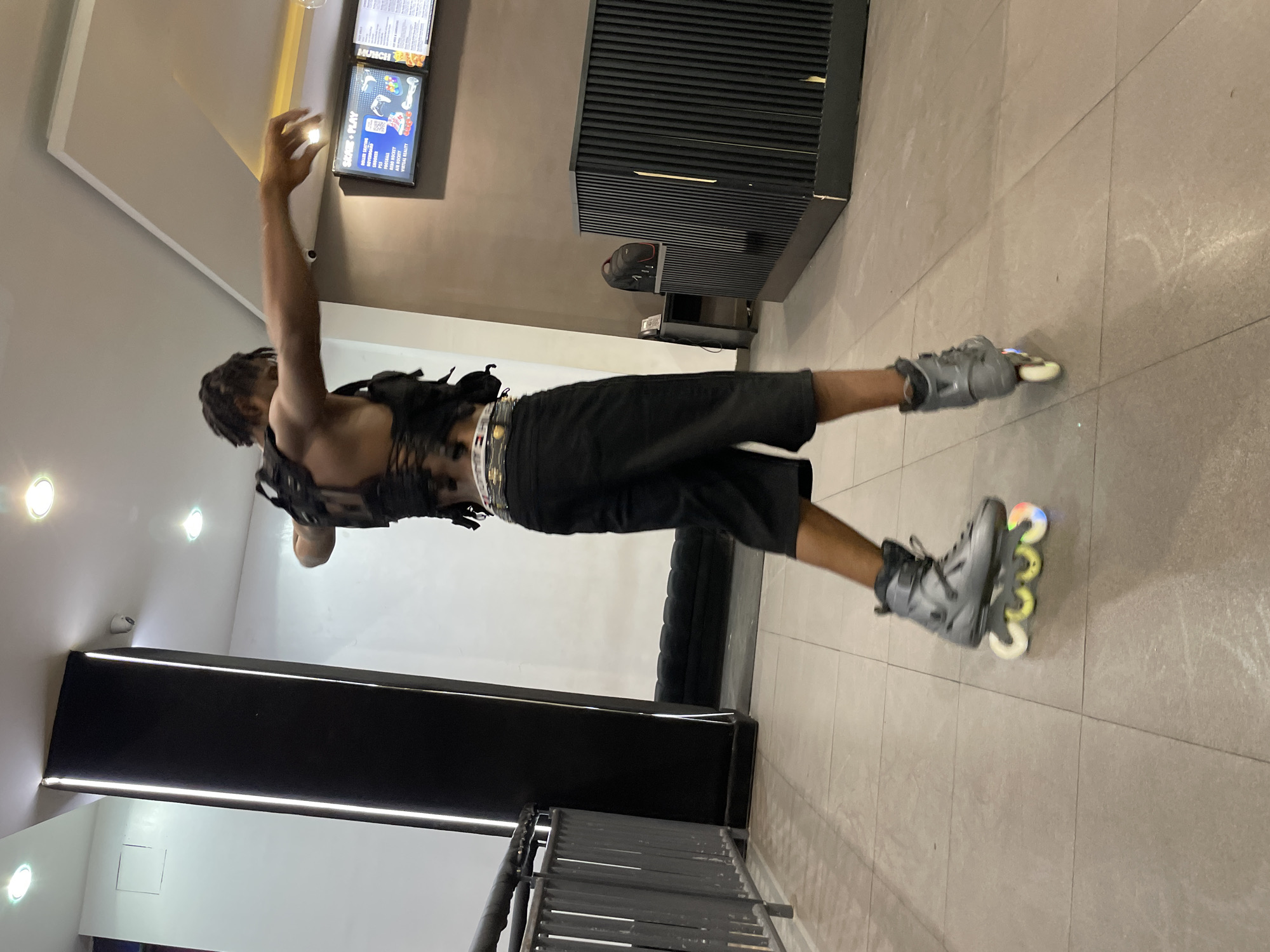 A young Black male is rollerblading indoors in what appears to be a recreational facility or entertainment venue. He is wearing black shorts, a black vest-like garment with lacing detail, and black rollerblades with illuminated wheels. The subject's back is to the camera as he skates away, his arms extended outwards. The floor is tiled, and the background features a dark pillar with vertical lighting, a white wall, and a counter area with a menu display. A black railing is visible on the left side of the image. The lighting suggests an indoor setting with overhead lights. The environment appears to be clean and modern.