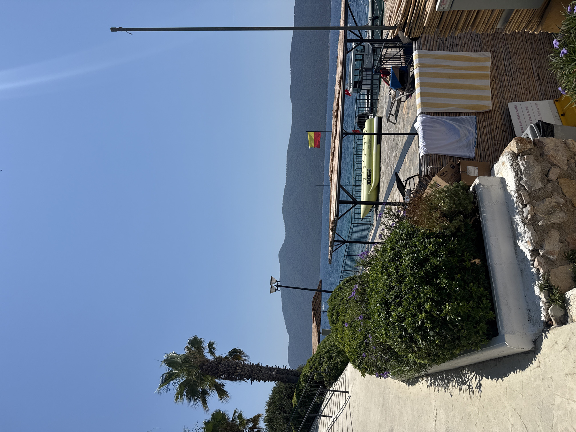 A sunny outdoor scene near Bodrum, Turkey, features a pathway on the left leading towards lush green bushes and flowering plants. Beyond the foliage, a calm blue sea stretches out to a distant, hazy mountain range under a clear sky. A wooden-roofed structure with a black metal frame sits by the water, supporting a yellow inflatable boat labeled "JOBE" and lounge chairs. A red and yellow flag flies from a pole near the structure. Two towels, one white and one striped yellow and white, hang on a bamboo-covered wall. In the foreground, a stone wall and a white planter box with more greenery are visible.