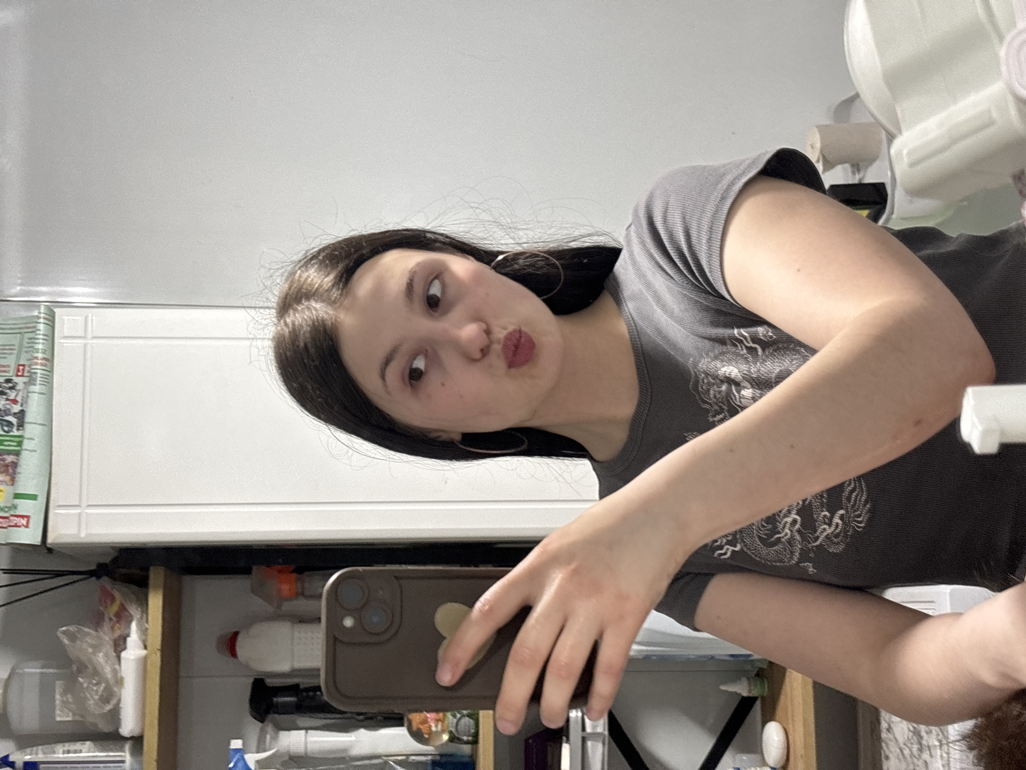 A young woman is taking a selfie in what appears to be a kitchen or utility area. She has dark hair, fair skin, and is wearing a gray t-shirt. Her lips are puckered in a kissing gesture. She is holding a brown iPhone with multiple lenses. Behind her, there is a white wall and cabinet. Below the cabinet is a shelf containing various cleaning supplies and other items. The exact location in the city of Фоки, Russia is not discernible from the image.