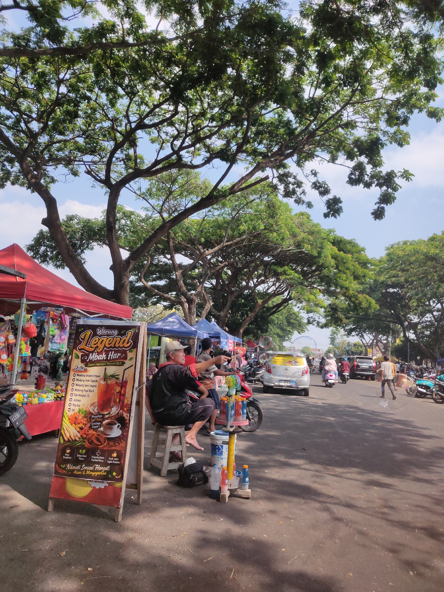 The image depicts a street scene in Ngasem, Indonesia, on a sunny day with a clear blue sky. Large trees with lush green foliage cast shadows on the asphalt road. In the foreground, a vendor is seated on a stool next to a sign for "Warung Legend Mbah Har," advertising various drinks like wedang empon, wedang jahe, wedang sereh, and coffee. The sign also features images of tea, coffee, and spices. To the left of the sign, a red tent displays an assortment of colorful toys and goods.
Several people are present in the scene. The vendor, wearing a black shirt and dark shorts, is looking to his right and appears to be interacting with someone out of frame. In the background, a silver car with a yellow sign on top is visible, along with several motorbikes and a few other cars. People are seen riding motorbikes and walking along the street. The overall atmosphere suggests a bustling market or roadside stall environment.