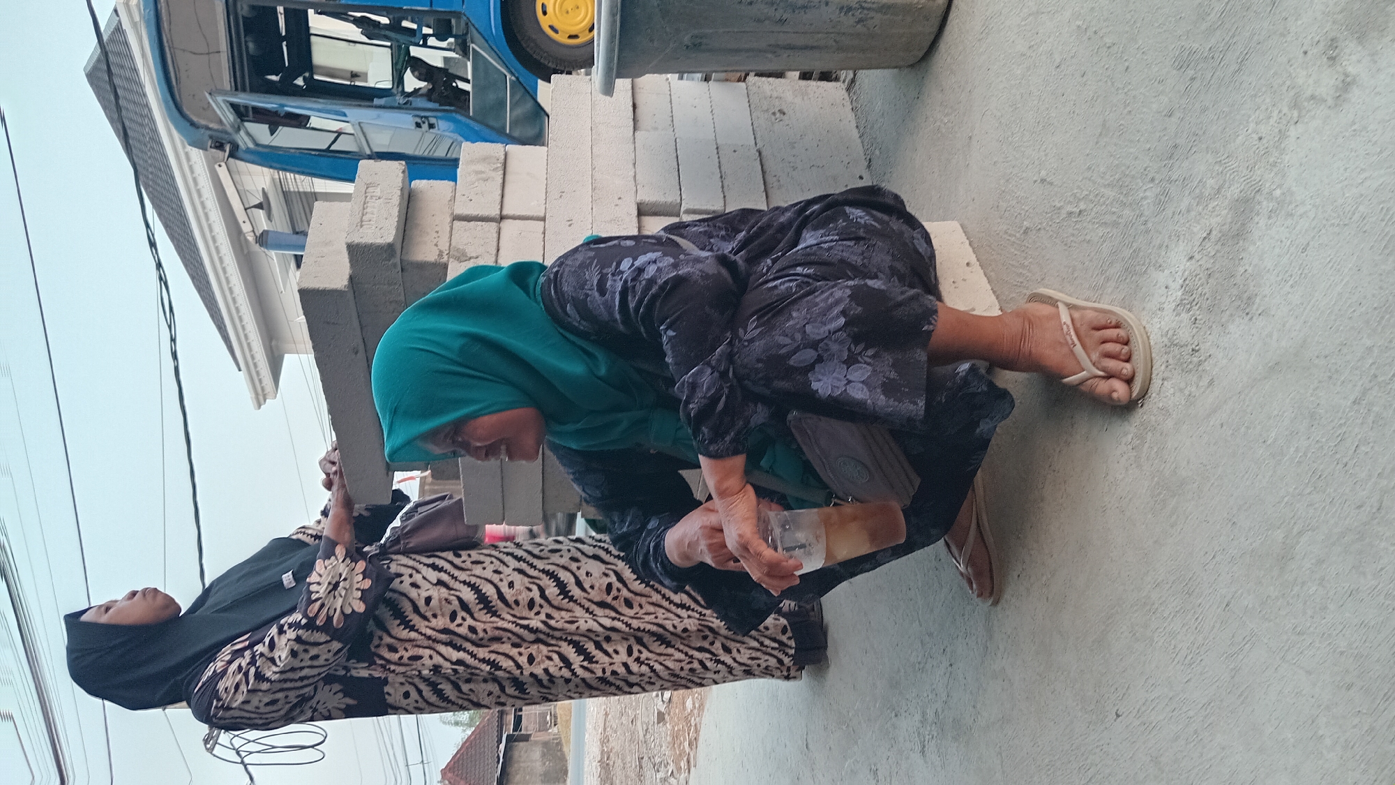 In Trembulrejo, Indonesia, two women are visible outdoors, in what appears to be a semi-urban or construction site area. The woman in the foreground is crouching and holding a plastic cup with a beverage. She wears a dark blue hijab and a patterned dark blue outfit. Her feet are bare except for light-colored flip-flops. Beside her is a small, grey cross-body bag. Behind her, to the left, stands another woman wearing a black hijab and a long, patterned black and white dress. She is leaning forward, resting her hands on a stack of concrete blocks.

The environment includes stacks of concrete blocks, suggesting ongoing construction or storage. In the background, a blue vehicle with an open door and a yellow wheel is partially visible. A grey trash bin is situated to the right of the scene. The ground is paved with concrete, and there is a slight incline. The sky above is overcast. The overall impression is a scene of daily life unfolding amidst a utilitarian or construction-related setting.