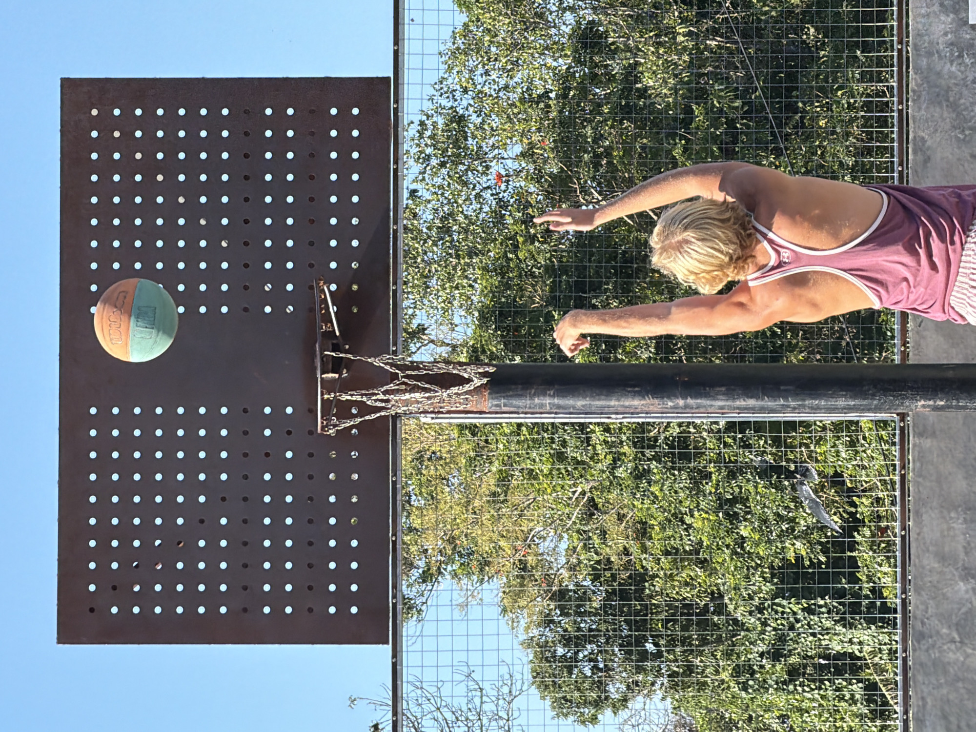 The image depicts a young person with blond hair, wearing a maroon tank top and striped shorts, in the act of shooting a basketball. The individual's arms are raised towards the hoop, and the basketball, a distinct orange and teal color with visible branding, is airborne just above the backboard. The basketball hoop, constructed from metal, features a chain net and is mounted on a dark pole. The backdrop consists of a rustic, perforated metal backboard set against a clear blue sky. In the background, lush green trees are visible behind a wire mesh fence, suggesting an outdoor setting, possibly a park or recreational area. The scene is captured from a low angle, emphasizing the action of the shot and the player's focus on the game.
