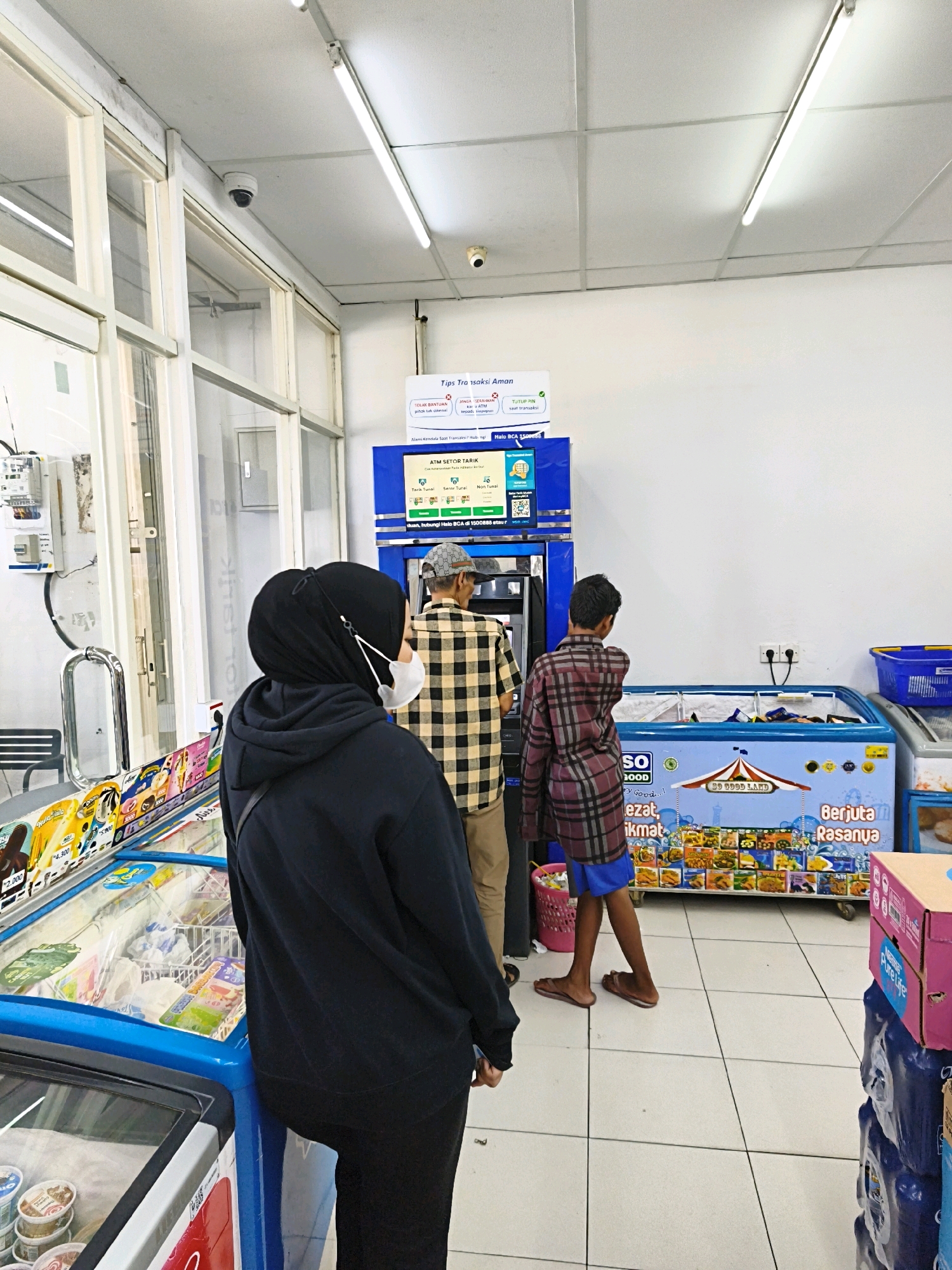 A medium shot captures the interior of a convenience store in Desa Donohudan, Indonesia, with a focus on an ATM machine and adjacent freezer units. In the foreground, a person wearing a black hijab and a black hoodie stands with their back to the camera, facing away from a glass-fronted freezer displaying various ice cream products. To the left of the ATM, a display of ice cream tubs and cones is visible, with pricing in Indonesian Rupiah.
Behind the person in the hijab, two individuals are interacting with the ATM. The person closer to the ATM is wearing a tan plaid shirt and a baseball cap. The second person, slightly further from the ATM and to its right, is a younger male wearing a maroon and black plaid shirt and blue shorts. They are both looking at the ATM screen.
The ATM is a blue unit with a screen displaying information and options. Above the ATM, a sign with Indonesian text and graphics provides transaction tips. To the right of the ATM, a large blue freezer unit is prominently displayed, featuring colorful graphics and the text "SO GOOD LAND" and "Berjuta Rasanya" (Millions of Flavors). Several product images are visible on the freezer door. Beyond this, other freezer units and shelves with products are partially visible.
The environment appears to be a brightly lit retail space with white tiled floors and white ceiling panels. Fluorescent lights are mounted on the ceiling. A security camera is visible on the upper left side of the image, mounted on the ceiling near a windowed section of the store. Electrical conduits and meters are present on a white wall section to the left of the ATM. A blue plastic basket is on top of a freezer unit on the far right. Stacks of boxed goods, including a pink box labeled "Pure Life", are visible in the lower right corner.