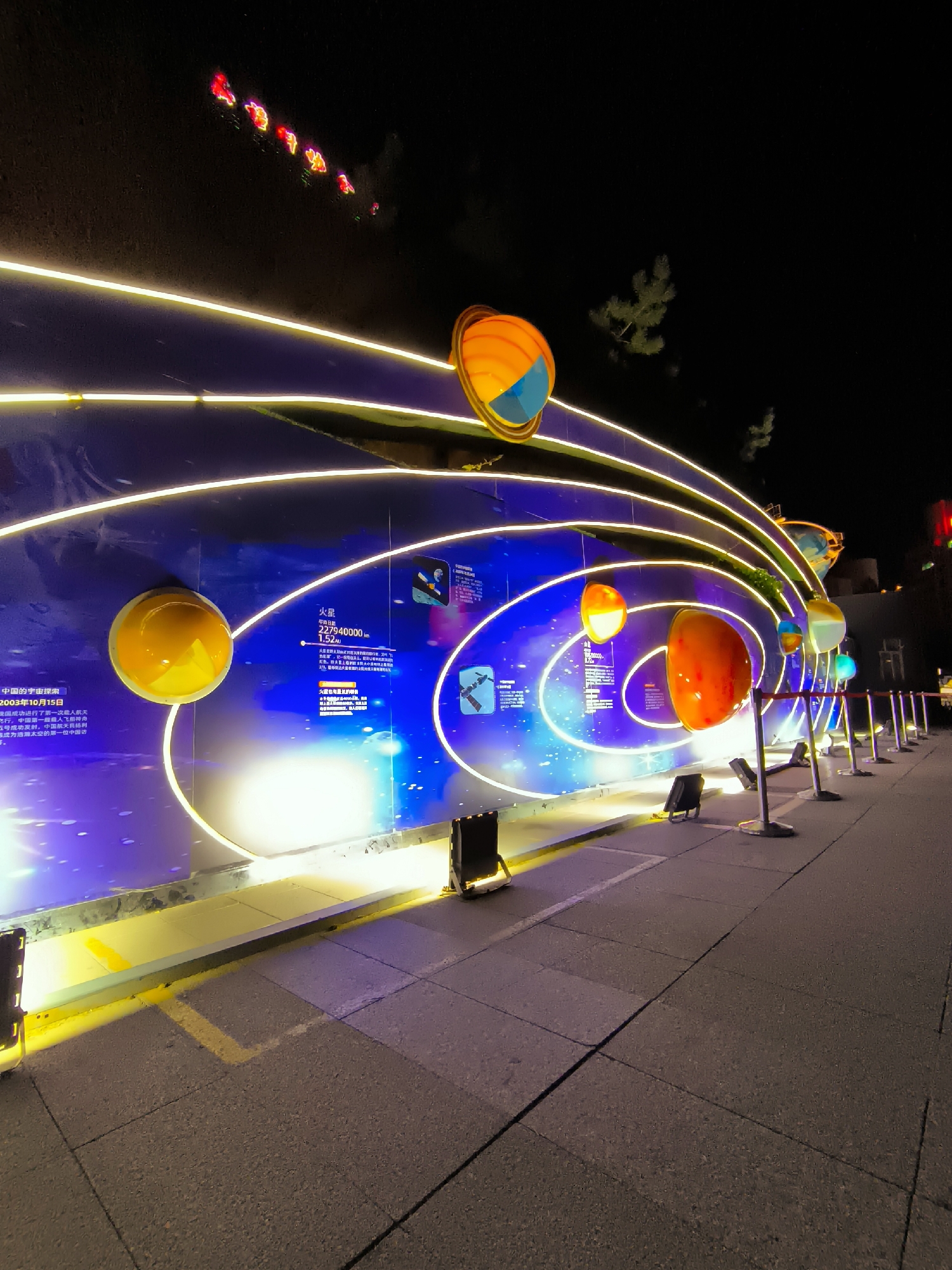 The image depicts a nighttime outdoor scene in Ürümqi, China, featuring a large, illuminated display that appears to be a model of the solar system. The display is set against a dark background, possibly a wall or building, which is lit with a vibrant blue and purple gradient, sprinkled with white dots resembling stars. Several large, spherical objects representing planets are suspended and illuminated, arranged in orbital paths marked by bright yellow neon-like lines. These lines curve and arc, creating a sense of movement and depth. The planets themselves are brightly colored, with shades of orange, yellow, and red prominently featured.
In the foreground, a paved walkway runs along the display. The walkway is made of square-shaped pavers and is illuminated by yellow and purple lighting cast from the display and some ground lights. Several small, black spotlights are positioned on the ground, aimed at the display. Metal stanchions with a reflective top are lined up along the edge of the walkway, separating it from the display area. In the upper portion of the image, a dark hill or elevated area is visible, with a string of red, lantern-like lights ascending its slope. A few scattered trees are also visible in the background. The overall impression is that of a public art installation or an educational exhibit, likely a part of a festival or event.