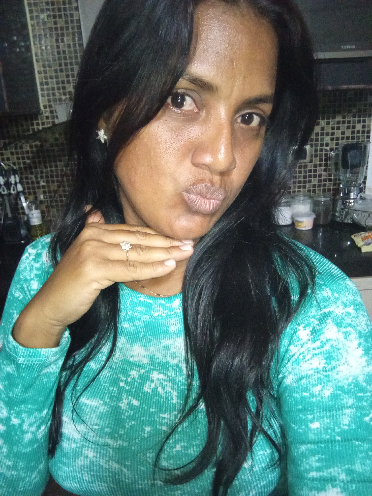 A woman is pictured from the chest up, facing the camera with her lips pursed. She has long, dark hair that partially obscures her face. She is wearing a teal, tie-dye patterned long-sleeved shirt and a ring with a flower-shaped stone on her right hand, which is touching her chin. The background appears to be a kitchen with tiled walls and various jars and appliances visible on a countertop. The lighting is dim.