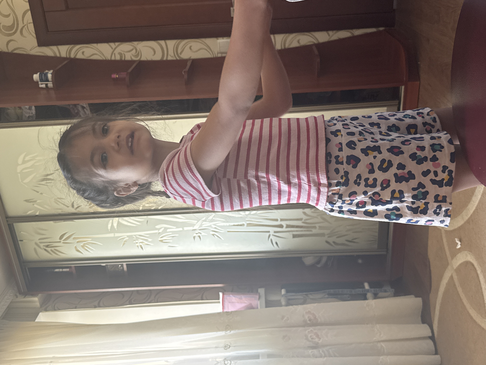 A young girl stands indoors, facing slightly right and looking towards the camera. She has dark hair styled with some strands framing her face and wears a small earring in her left ear. Her attire consists of a ribbed t-shirt with horizontal pink and white stripes and white shorts featuring a multi-colored leopard print in blue, pink, yellow, and black. Her mouth is slightly ajar, and her right arm extends out of frame to the right.
The background includes a light-colored, sheer curtain with subtle patterns on the far left, partially obscuring a window and a pink container on a sill. Adjacent to the curtain, a dark wooden cabinet or wardrobe with a frosted glass sliding door displays a bamboo leaf design. To the right of the girl, a dark reddish-brown wooden furniture piece, possibly a shelf unit, contains a white bottle and a smaller white container on its top shelf, and a small pink object on a lower shelf. The wall behind this furniture is covered with patterned wallpaper featuring a light beige/gold and darker beige/brown swirling design. The floor is carpeted in a light brown/beige hue with a lighter swirling pattern, and a small, crumpled white object rests on it. A dark red or maroon circular object, possibly a stool or cushion, is partially visible in the bottom right foreground. The scene is illuminated by natural light entering from the left. Location is Unknown, Unknown.