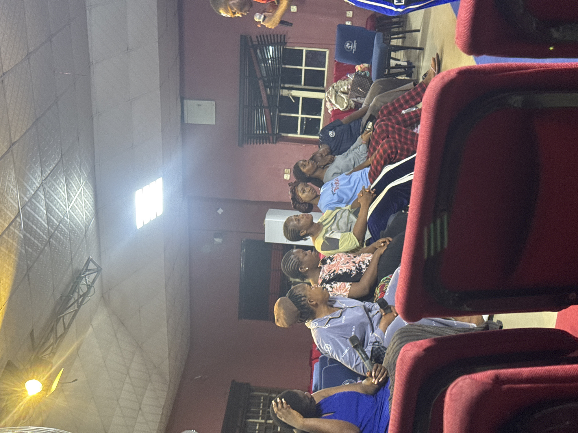 A group of people are seated in rows of red chairs in what appears to be a hall or auditorium in Onitsha, Nigeria. Most of the attendees are women. A speaker stands at the front of the room, holding a microphone. A window can be seen in the background, and the room is illuminated by artificial light. The ceiling has an elaborate grid pattern.
