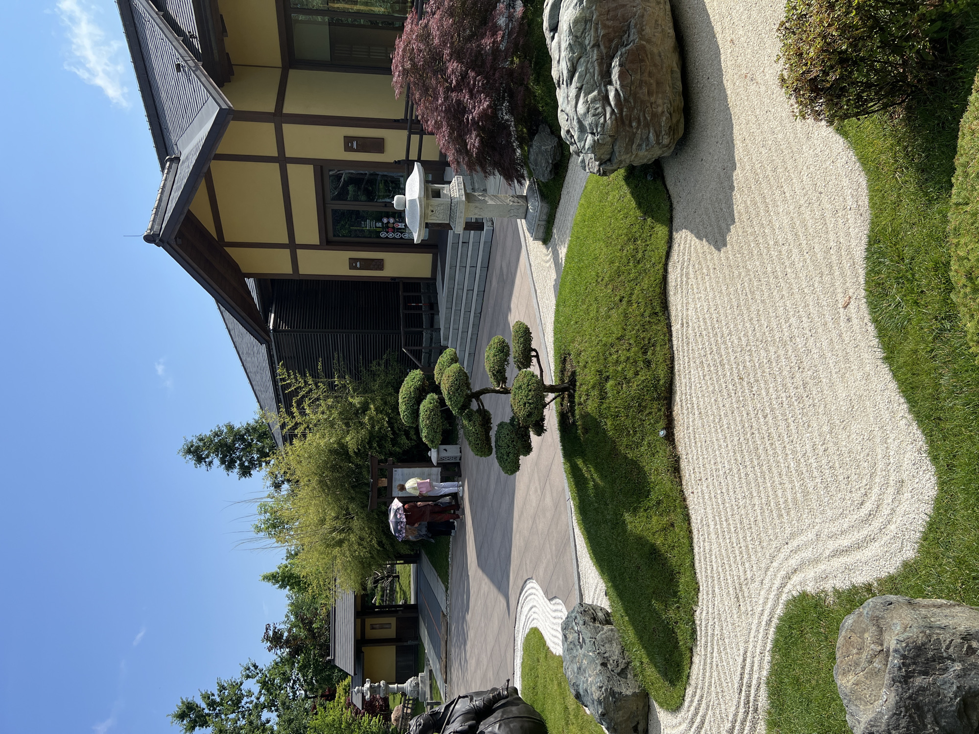 This image depicts a Zen garden scene at the "Garden of Eden" park in Krasnodar, Russia. The foreground features a meticulously raked white gravel area with sweeping curves, bordered by vibrant green grass. Several large, natural rocks are strategically placed within the garden. A meticulously pruned bonsai tree stands on a grassy mound in the center of the frame. In the midground, a stone lantern and a Japanese maple tree with deep red foliage are visible. Behind the garden elements, a building with traditional Japanese architectural features, including a yellow facade and dark tiled roof, serves as the backdrop. Several individuals are standing near the entrance of the building, some holding umbrellas. Lush greenery, including bamboo, surrounds the building and garden. The sky is clear and blue.