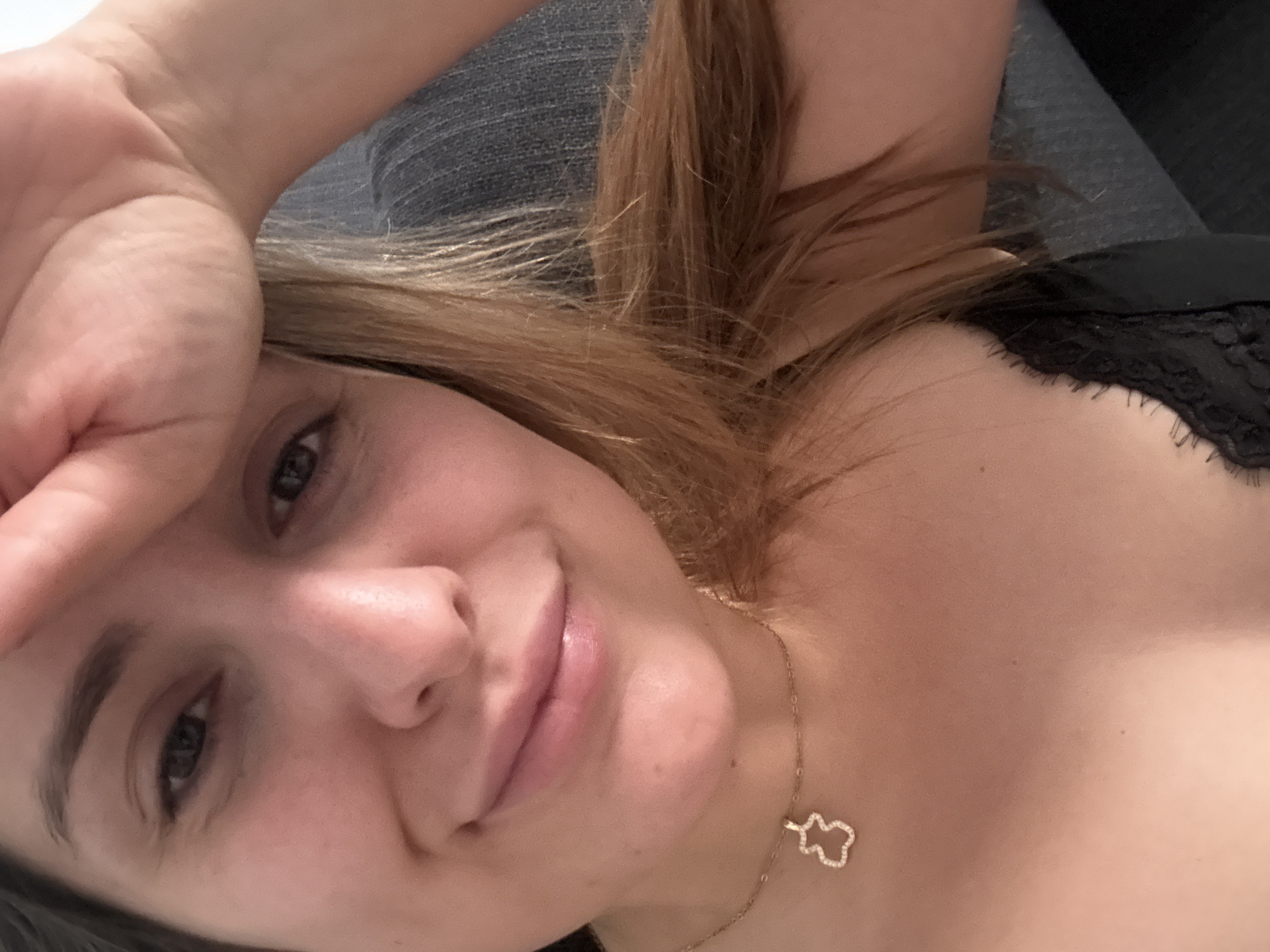 A selfie shows a young woman with fair skin and light brown hair. She is lying down, looking directly at the camera with a slight smile. Her right hand is raised to her forehead. She wears a gold necklace with a bear-shaped pendant. The background is blurred, but suggests a couch with gray and black fabrics. There is no information to determine if the location is in Barcelona, Spain.