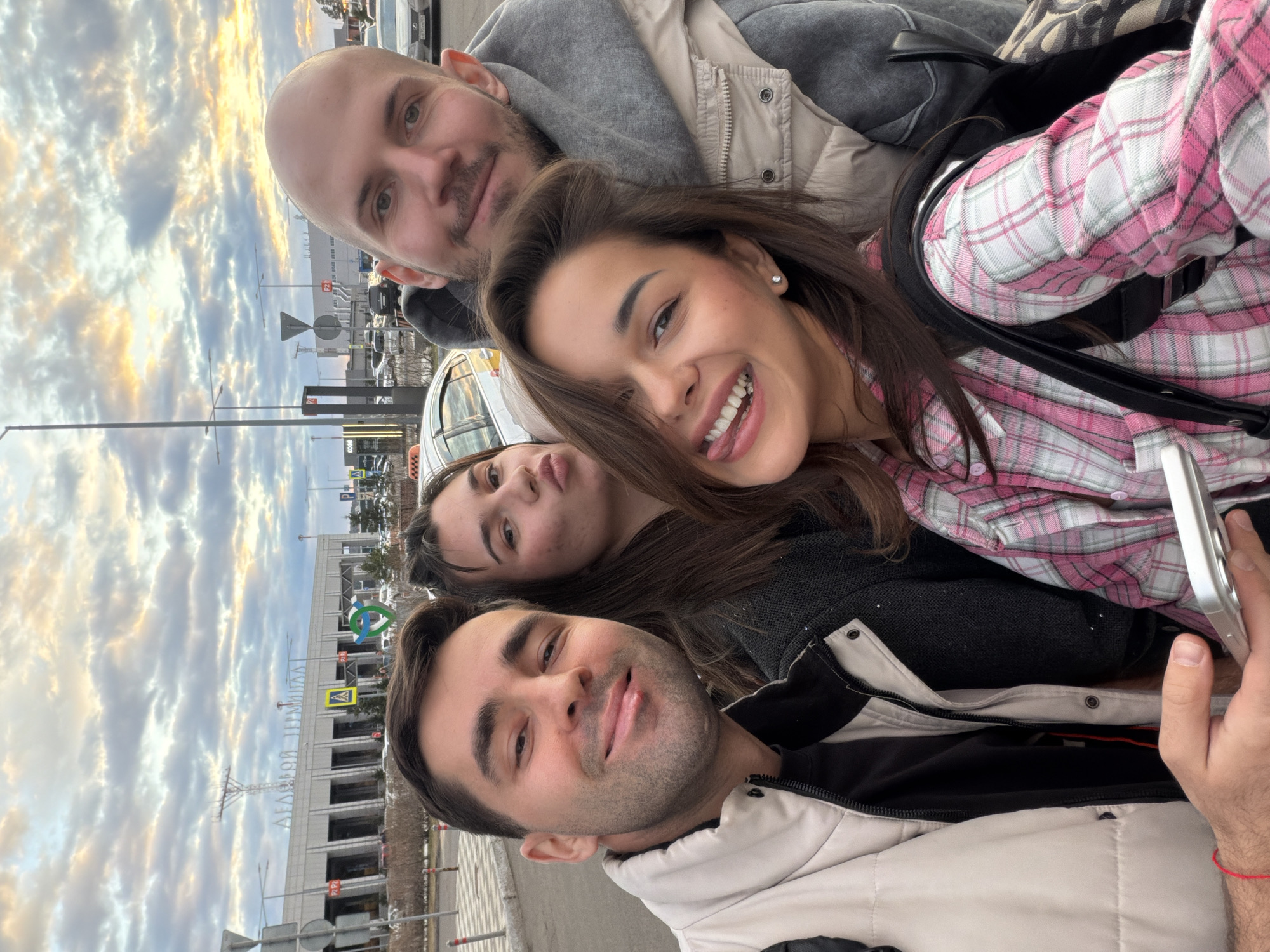 Four people are posing for a selfie outside the Yemelyanovo International Airport in Yemelyanovo, Russia. The airport terminal is visible in the background, with the name "АГАФИОН ТЕРМИНАЛ" prominently displayed. Above that, a green and blue logo can be observed. One person has a pink and white plaid shirt. The person taking the photo is wearing a light tan jacket. The sky is cloudy.