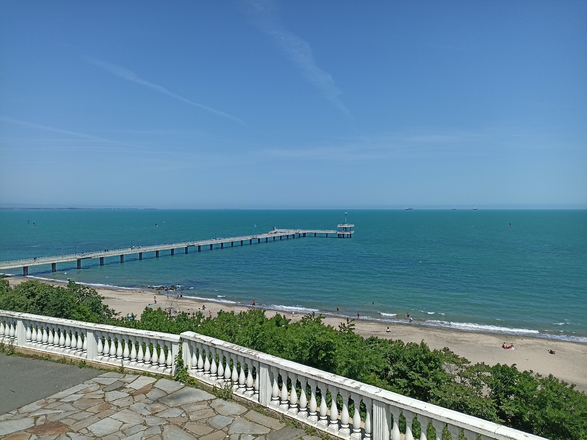 The image presents an elevated view of the Burgas Pier and beach area along the Black Sea coast in Burgas, Bulgaria, under a clear blue sky.
In the immediate foreground, a pathway paved with irregular, light-colored flagstones of varying sizes occupies the bottom left. A white, ornate balustrade, composed of numerous identical balusters, extends diagonally from the bottom-left corner towards the mid-right. Minor green vegetation grows along the base of the balustrade, which exhibits some weathering.
Below the balustrade, a dense strip of green deciduous trees and bushes lines the top edge of a sandy beach. The beach is populated by several individuals and small groups. Towards the left, a metal climbing frame or similar recreational structure stands on the sand, with a few people nearby. Further right on the beach, two individuals are seen lying on a red blanket. Additional people are dispersed along the shoreline, some wading in the shallow, turquoise-blue water where small waves are visible.
A long, light-colored pier, supported by a series of evenly spaced pillars, extends directly from the beach into the sea. Multiple individuals are visible walking along the pier. At its end, a prominent circular structure with multiple levels and an observation platform is present.
The sea, a consistent turquoise-blue hue, covers the majority of the background. On the water's surface, particularly towards the left of the pier, several small craft with sails, indicative of windsurfers or kite surfers, are in motion. On the distant horizon, towards the right, three distinct large vessels, possibly cargo ships, are visible. A faint landmass is discernible on the far left horizon.
The sky is a uniform, bright blue, largely clear, save for two faint, linear contrails that traverse the upper portion of the frame.