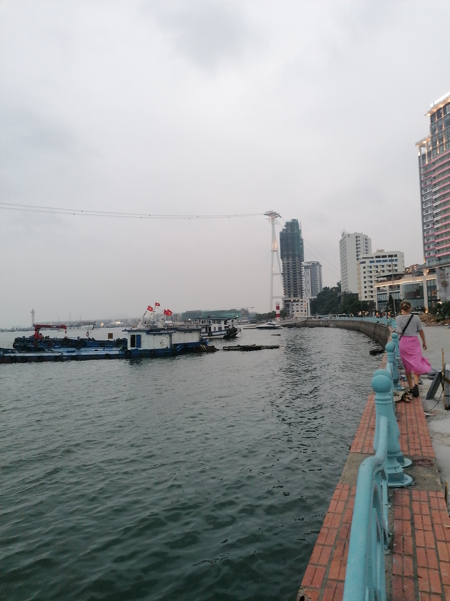 A person wearing a gray shirt and a pink skirt walks on a brick walkway alongside a body of water. A light-blue railing borders the walkway. In the water, there are several boats, some adorned with red flags. Across the water are buildings and a tower under construction. The sky is overcast. The location is Bai Chay Ward, Vietnam, based on the architecture and general environment.
