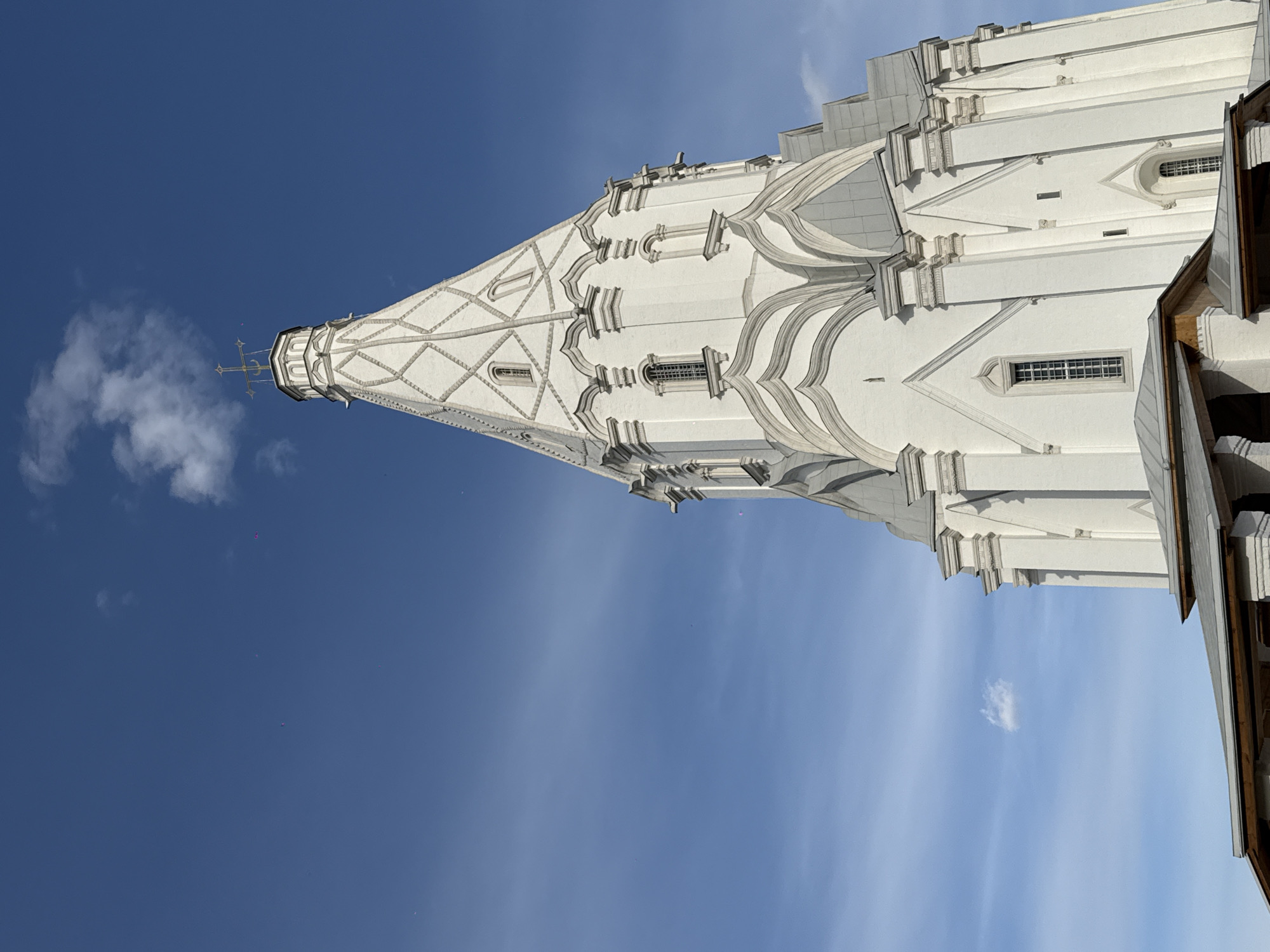 The image captures a portion of the Church of the Ascension in Kolomenskoye, Moscow, Russia. The white building features a tall, tent-like spire with decorative elements and windows. The spire is topped with a cross. The sky is blue with some light clouds. A small portion of a building with wooden trim is visible on the right side of the frame.
