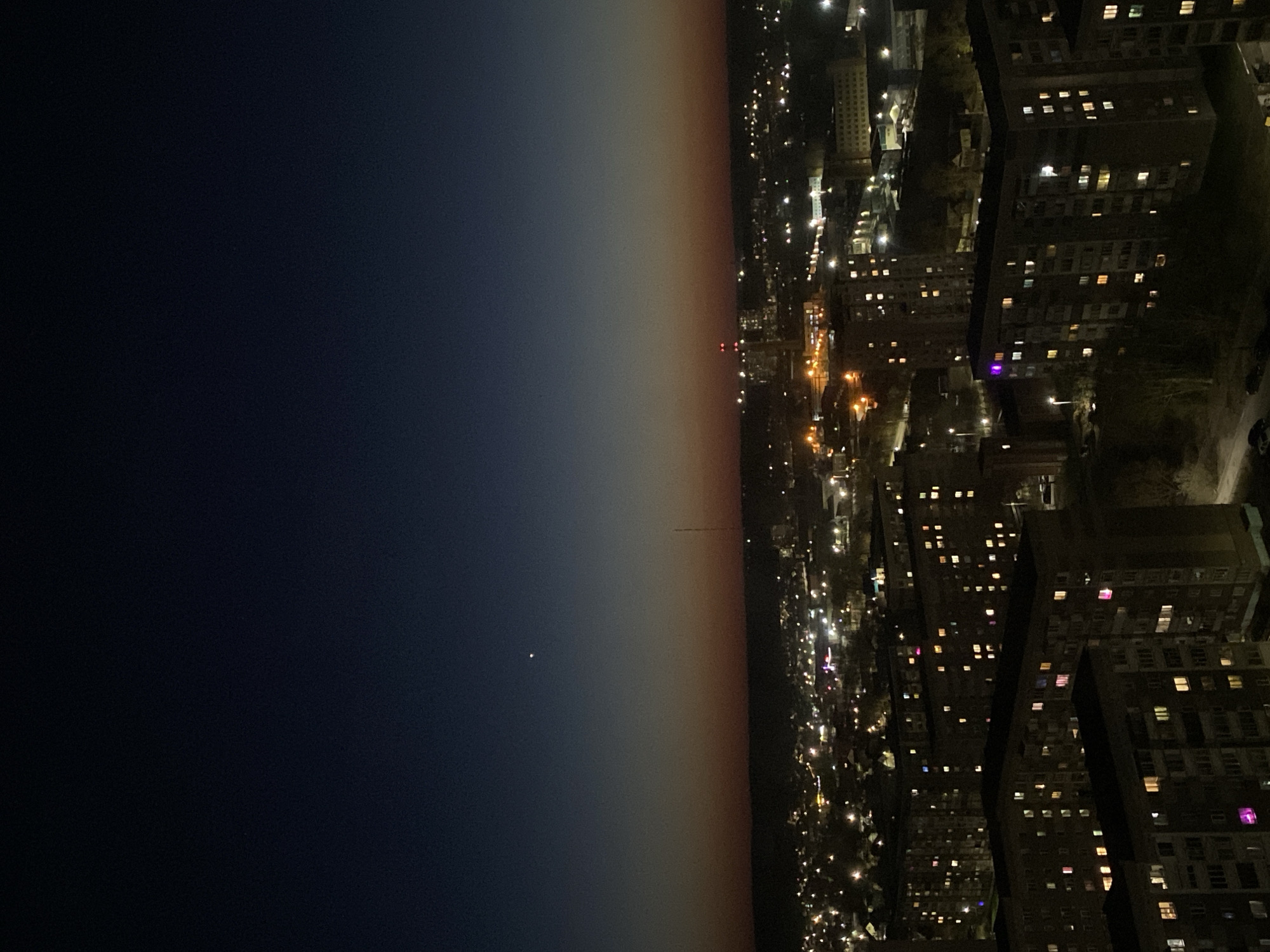 The image shows a night view of Izhevsk, Russia, with a stark contrast between the dark sky and the illuminated cityscape. The sky transitions from deep blue to a pale orange gradient near the horizon, where a small, bright celestial object, likely a star or planet, is visible. The city is represented by dense clusters of lit buildings, primarily residential blocks with numerous windows displaying lights. The streetlights form long, winding paths through the urban fabric, hinting at the layout of roads and streets below. The image lacks any visible people.