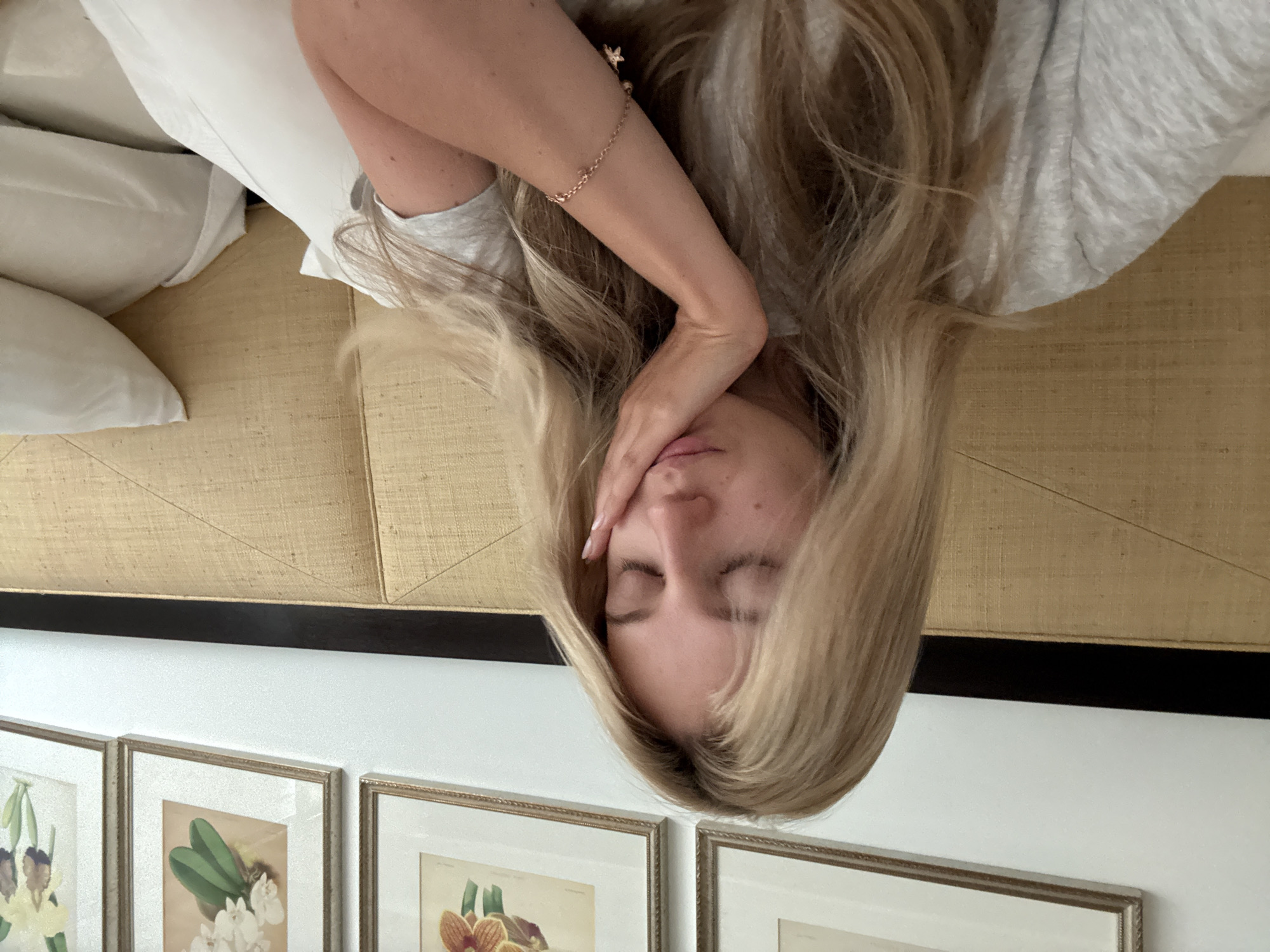 A medium shot shows a fair-skinned young woman with long, straight blonde hair resting her cheek on her hand. Her eyes are closed, and her lips are slightly parted. She is wearing a light gray t-shirt and a delicate gold bracelet with star charms on her left wrist. Behind her is a textured gold headboard of a bed, and to her right, white pillows are visible. The background includes a white wall with framed artwork depicting flowers. The scene appears to be set in a hotel room in Mexico City, Mexico.