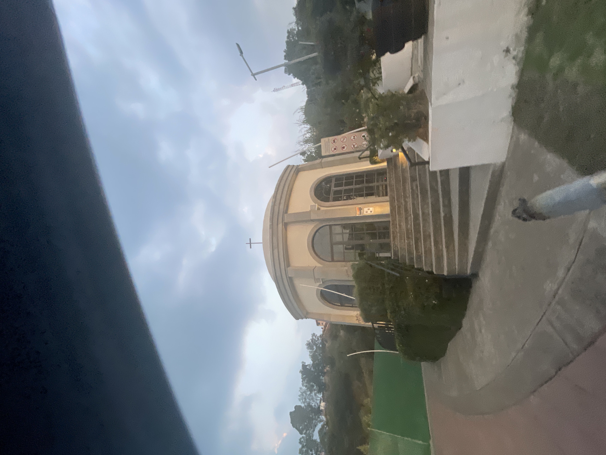 The image shows the rotunda-shaped Capilla San José del Cubo church, located in Huixquilucan de Degollado, Mexico. The church, constructed with light-colored stone, features arched windows and a cross at the apex of its dome. A stairway leading up to the church entrance is visible, along with some signage and landscaping around the building. The sky is overcast. A partial view of a sports field is seen on the left side of the church.