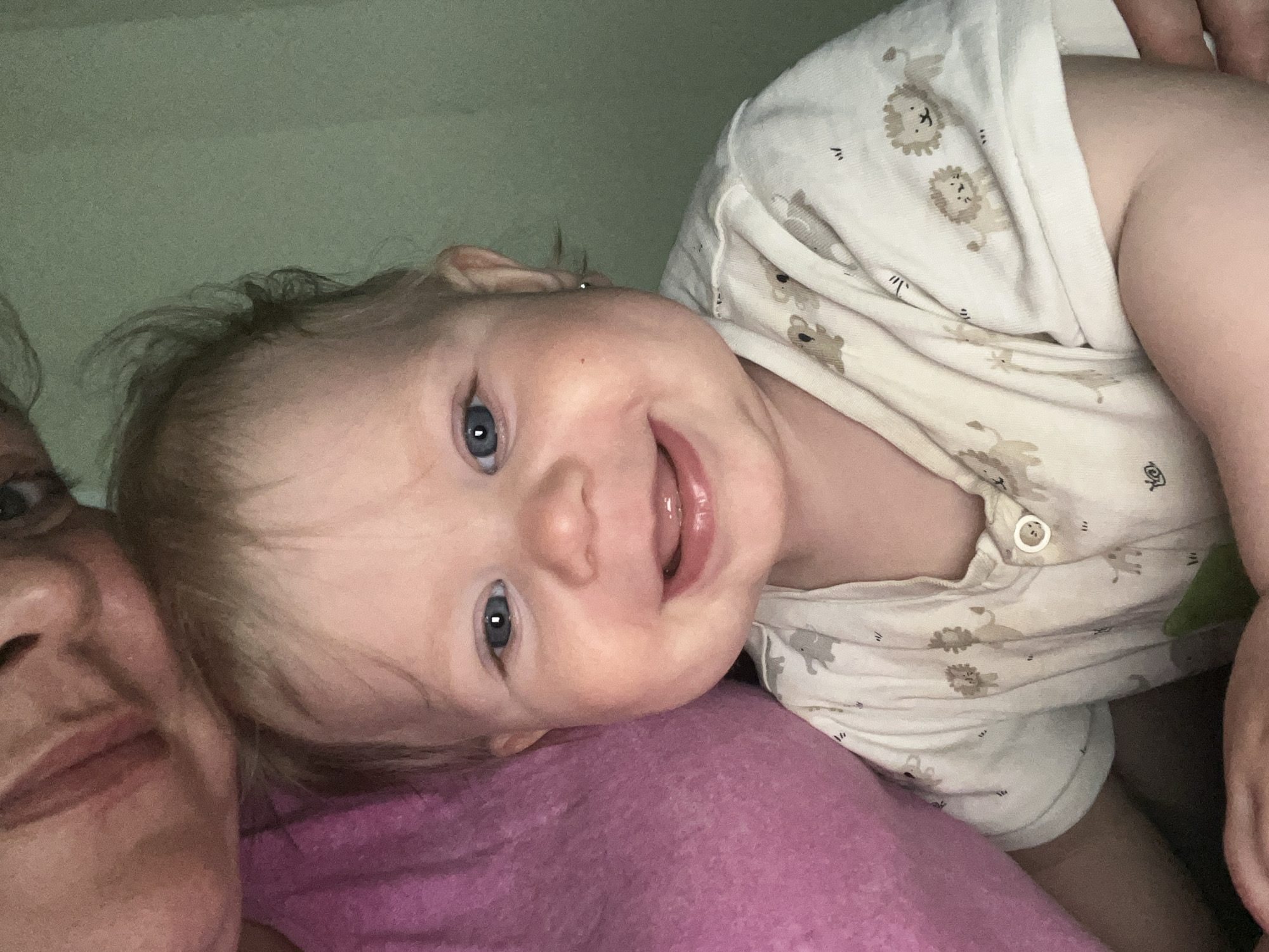 An intimate, close-up photograph features a baby and a partially visible adult, both appearing to be lying down indoors. The baby, positioned prominently, has fair skin, light-colored hair, and bright blue eyes, looking directly at the viewer with a wide, joyful smile that shows off gums and a pink tongue. The baby is dressed in a light-colored onesie or pajamas patterned with small, repetitive designs of lions and giraffes.
On the lower left side of the frame, the left profile of an adult's face is visible, showing the mouth, chin, nose, and part of one eye, indicating close proximity to the baby. Both individuals are resting on a soft, textured pink or magenta blanket or fabric. The background consists of a plain, light green wall or surface, suggesting an indoor setting such as a bedroom. The lighting is soft and even, without harsh shadows, which implies a relaxed atmosphere, possibly in the evening or a dimly lit room. The overall impression is one of warmth, affection, and happiness shared between the adult and the smiling baby. No discernible text is visible in the image.