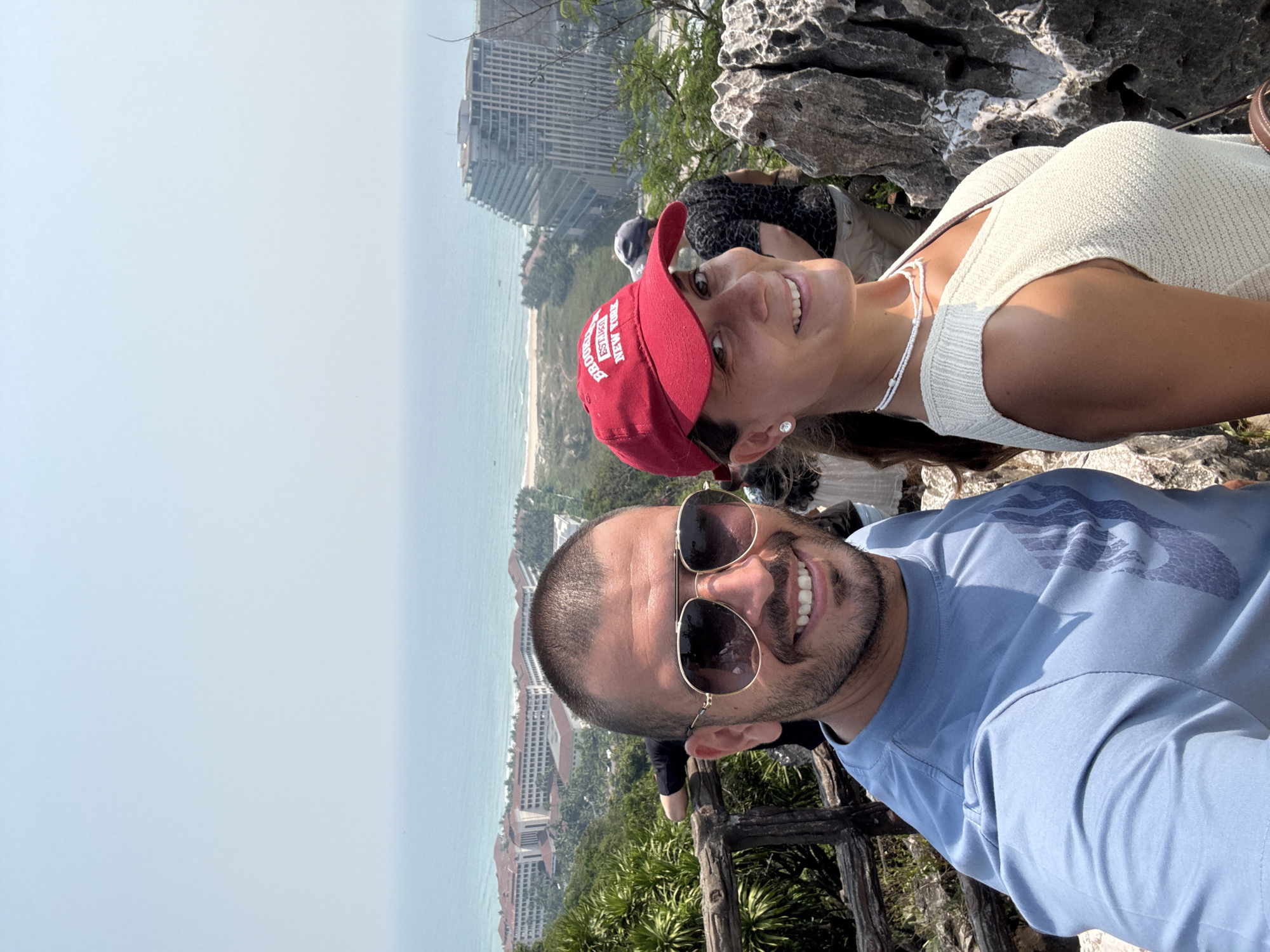 A selfie depicts a man and a woman at a high vantage point overlooking Đà Nẵng, Vietnam. The man, in the foreground, is wearing sunglasses and a blue t-shirt. The woman, slightly behind him, is wearing a white top and a red baseball cap. A cityscape and ocean are visible in the background. Vegetation is present in the foreground. The setting appears to be a rocky outcrop.