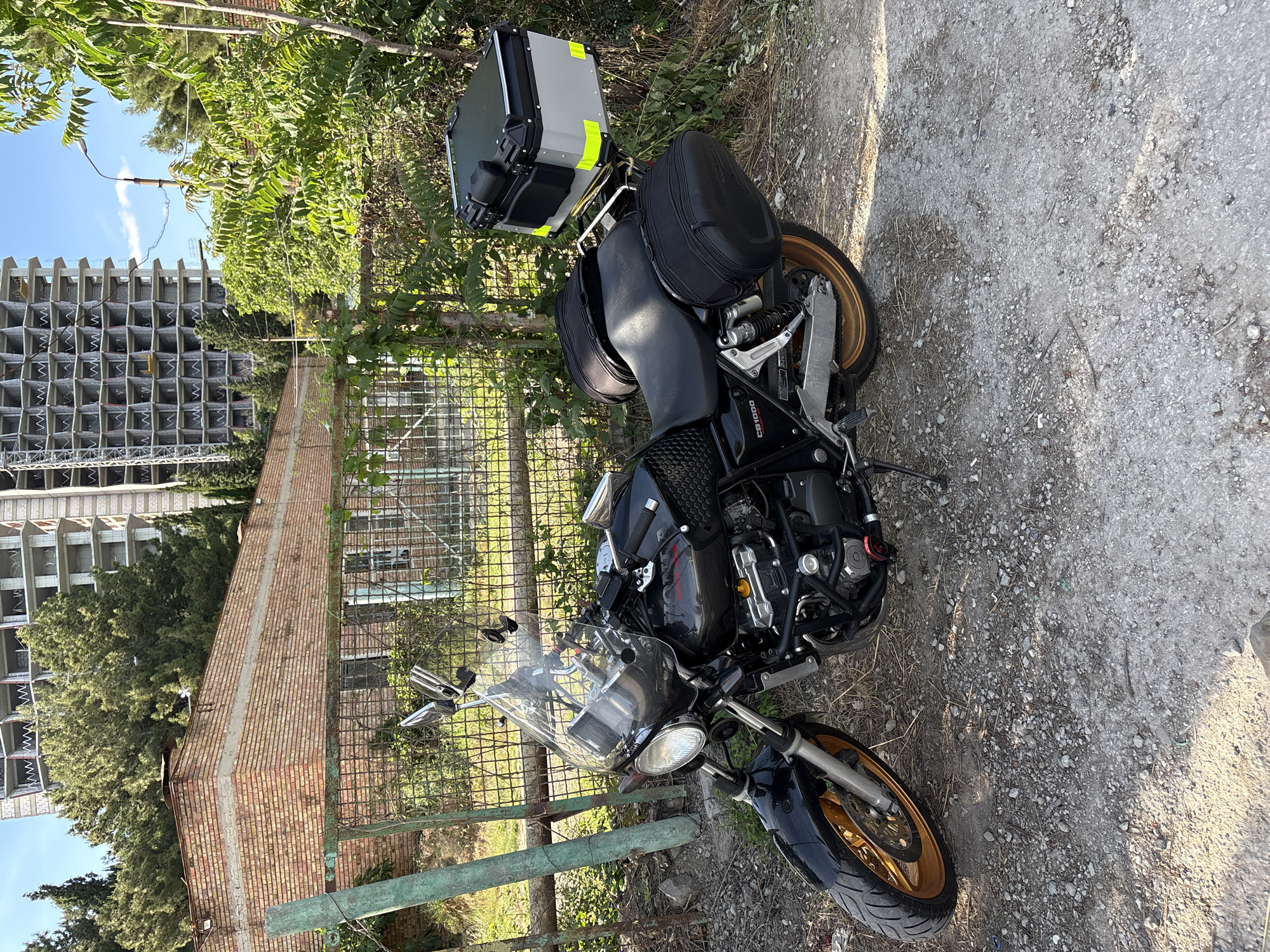 This is an eye-level, slightly elevated, outdoor, long shot of a parked motorcycle in a partially developed area. The motorcycle is black with gold rims and is equipped with saddlebags and a top case, suggesting it is set up for touring. It is parked on a gravel and dirt path next to a green metal fence. Behind the fence, there is a brick building with a damaged roof. Further in the background, a large, unfinished concrete building with visible structural elements stands under a clear blue sky with a few wispy clouds. Lush green trees and foliage surround the scene. The lighting suggests it is daytime, likely sunny. There is no visible text on the motorcycle or in the immediate surroundings. The overall impression is one of a temporary stop in a location undergoing construction or redevelopment. The scene is static, with no activity taking place beyond the motorcycle being parked.