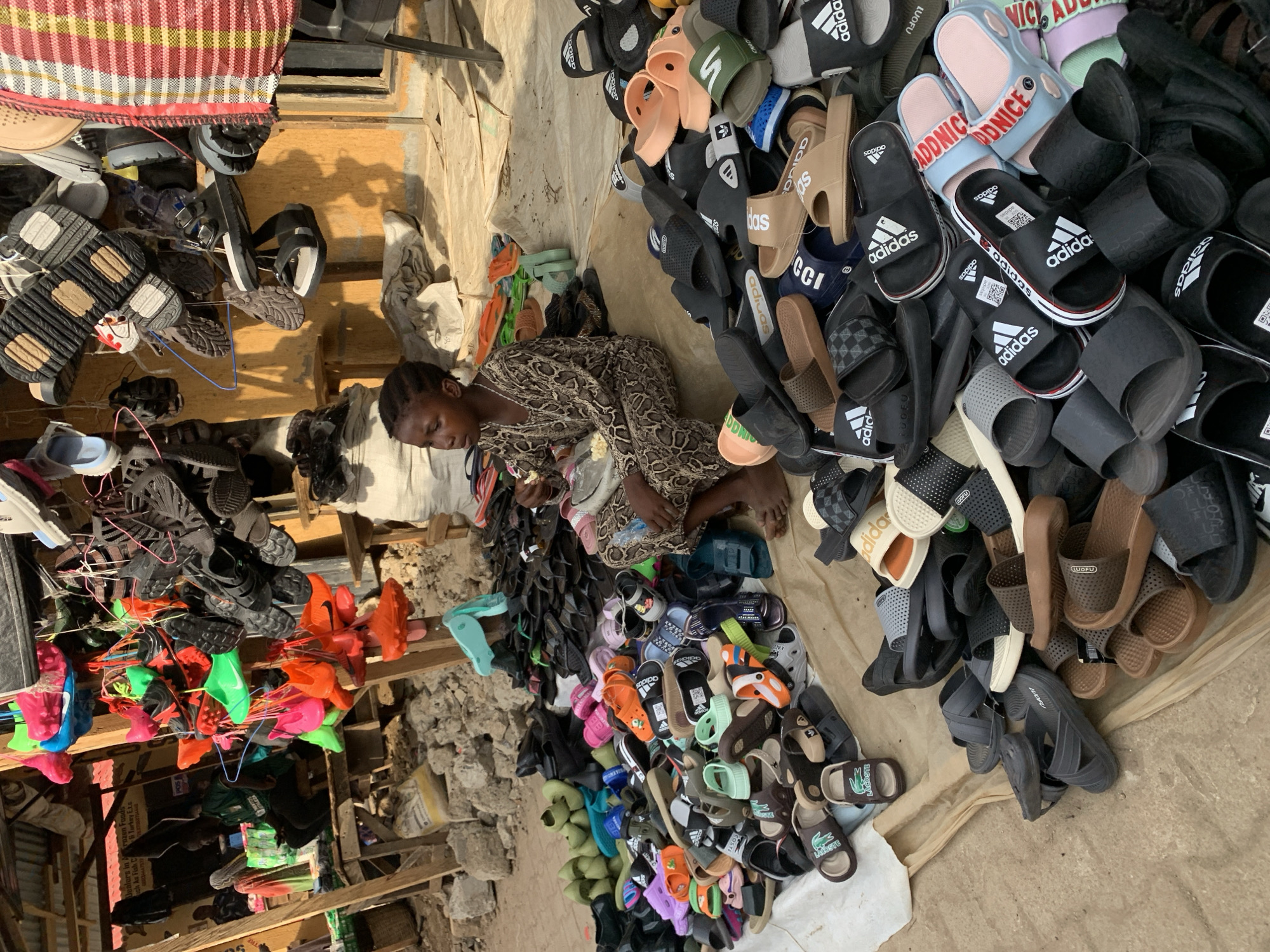 The scene depicts a woman surrounded by a large assortment of footwear, likely in an outdoor market setting in Kafanchan B, Nigeria. She is sitting on the ground, potentially examining or sorting through the sandals. Piles of sandals, a variety of styles and colors, fill the foreground. Some bear brand names, such as "Adidas" and "Lacoste." Additional footwear, including athletic shoes, hang from a wooden structure in the background. The environment appears to be a modest commercial area, possibly with a retail establishment nearby.
