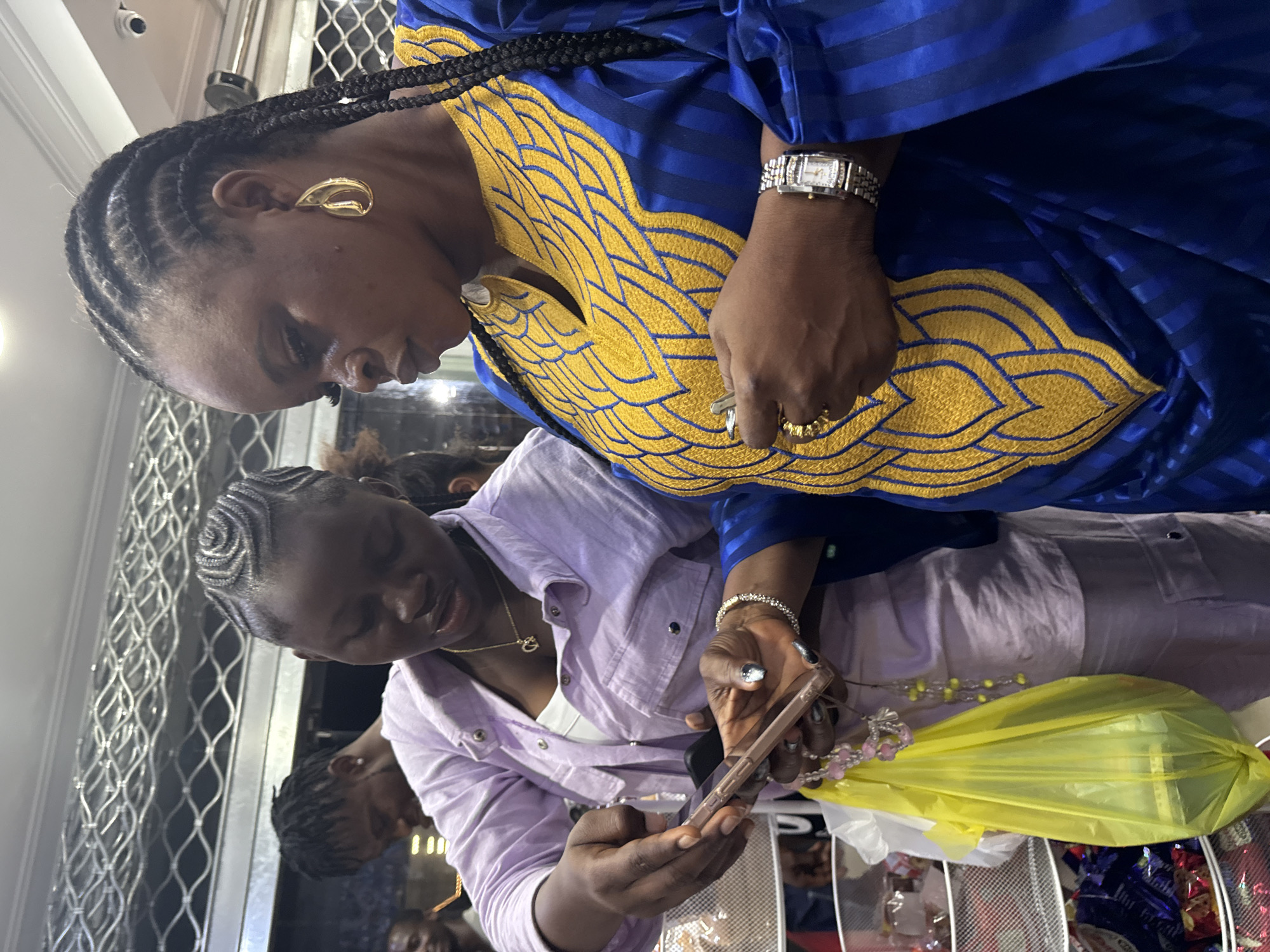 This image depicts a scene in the city of Ado, Nigeria, within an indoor commercial establishment, likely a shop or market stall. In the foreground, two women of dark complexion are engaged in an interaction centered around a smartphone.
The woman on the left, positioned slightly forward, wears a light lavender button-up shirt over a white top and matching light-colored pants. Her hair is styled in intricate cornrows forming concentric spiral patterns on her scalp. She holds a light-colored smartphone in her left hand, while her right index finger points directly at its screen. A gold-colored necklace with a "C" or heart-shaped pendant is visible, along with a multi-strand pink and white beaded accessory dangling from her right hand, alongside a yellow plastic bag containing an indistinct green object. Her fingernails are painted with a dark tip over a lighter base.
The woman on the right wears a distinctive royal blue traditional garment featuring prominent gold-colored embroidery in stylized feather or leaf patterns across the chest and V-neckline. Her hair is styled in braids, with a large gold-colored teardrop-shaped earring visible in her right ear. A silver-toned wrist watch with a rectangular, stone-embellished face is on her left wrist, and a gold-colored ring is on her left ring finger. She holds a small metallic object in her left hand, and her gaze is also directed at the smartphone screen.
In the mid-ground behind the woman on the left, a man with dreadlocks or braids is partially visible, looking downwards. Further to the left, part of another man's face, smiling, can be seen. The background reveals white metal mesh shelving units filled with various packaged goods, consistent with a retail environment. A metallic grid-like structure is present on the upper wall or ceiling, and a small black security camera is mounted on the upper right wall. The scene is illuminated by artificial lighting.