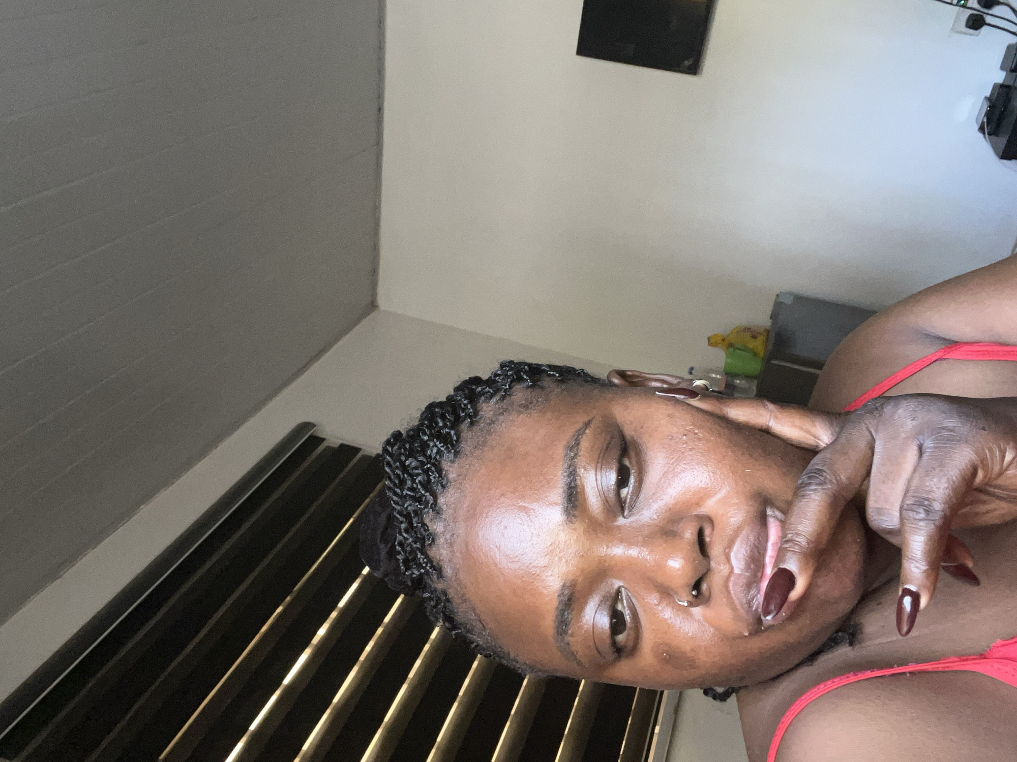 An individual with dark skin and braided hair is shown in a close-up selfie. They are wearing a red top and have their finger near their mouth. The background includes blinds, a white wall, and some objects on a shelf. The scene is lit by natural light.