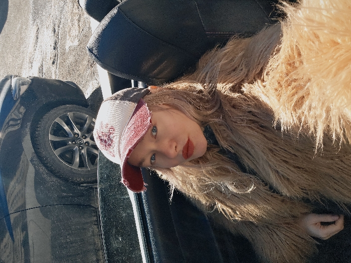 A young adult female is depicted seated in the passenger side of an open-top vehicle, facing the camera. She has blonde, wavy hair and wears a distressed white and maroon baseball cap, red lipstick, and a shaggy, light brown faux fur coat. Her left hand rests on her lap. In the background, a dark-colored passenger vehicle, possibly a sedan or SUV, is partially visible, its lower body and wheels soiled with dirt or grime. The vehicle is parked on an unpaved or dirty ground surface that exhibits patches of reflective material, possibly water or dirty ice/snow. The scene is outdoors and brightly lit by sunlight, casting distinct shadows and highlights. The specific location within Saint Petersburg, Russia, is not discernible from the visual information provided, but it is an outdoor setting.