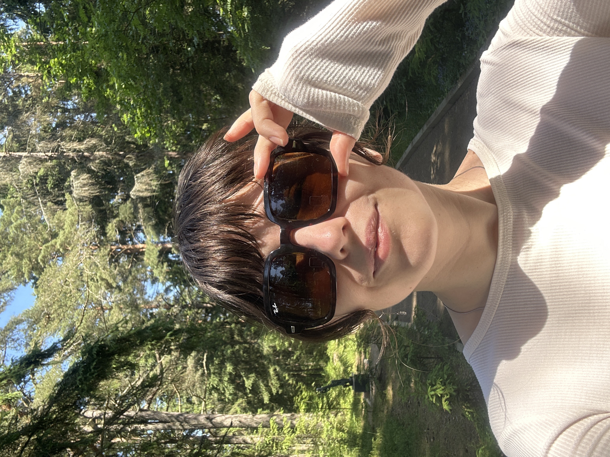 The image shows a close-up of a person wearing large, dark sunglasses with the Ray-Ban logo visible. The person has dark hair with bangs and is wearing a light-colored, ribbed long-sleeved shirt. Their right hand is raised, holding the sunglasses. The background is a sunlit forest with tall trees and a paved path. A small statue of a person is visible to the left of the path. The lighting suggests it is daytime. The image was taken in the city of Shepelyovo, Russia.