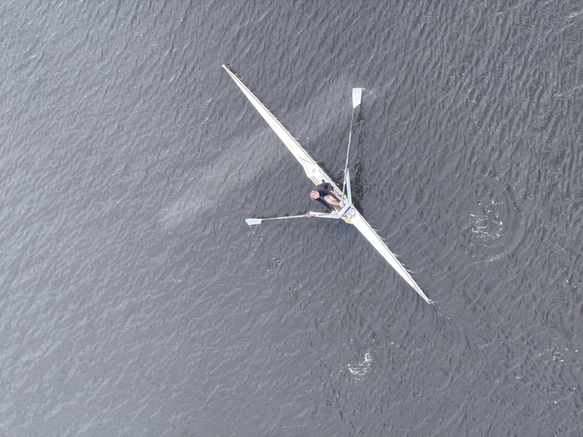 An aerial view shows a single scull rowing boat on a body of water, likely the Volga or Oka River in Nizhny Novgorod, Russia, given the landscape. The boat is pointed diagonally across the frame from upper right to lower left. A lone rower is visible in the boat, actively engaged in rowing. Ripples and disturbances in the water indicate the boat's recent passage and activity. The water surface appears textured, suggesting a slight breeze or current.