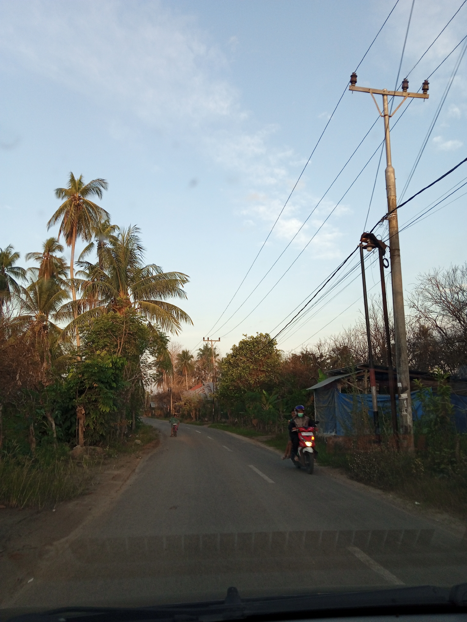 The image shows a road scene in Kubu, Indonesia. The view is from inside a vehicle, looking out onto a paved road that curves slightly to the left. Two motorcycles are visible on the road, one closer to the camera with two occupants and another further away. A utility pole with power lines is prominent on the right side of the road. Lush vegetation, including palm trees, lines the road. A partially visible structure with blue tarps can be seen on the right side. The sky is light blue with scattered clouds, suggesting either morning or late afternoon light.