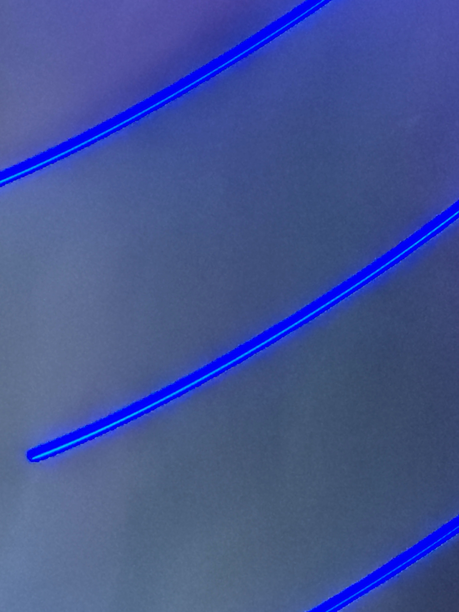 The image shows a close-up of a surface with three diagonal blue neon lights. The lights are parallel and appear to be embedded in or mounted on the surface, which is a muted gray color. The lighting is even, and there are no visible shadows or reflections. The image does not depict any identifiable landmarks or elements that would allow for a specific location within Bangkok, Thailand, to be determined. It is a purely abstract and minimalist composition focusing on the geometric lines of the neon lights.