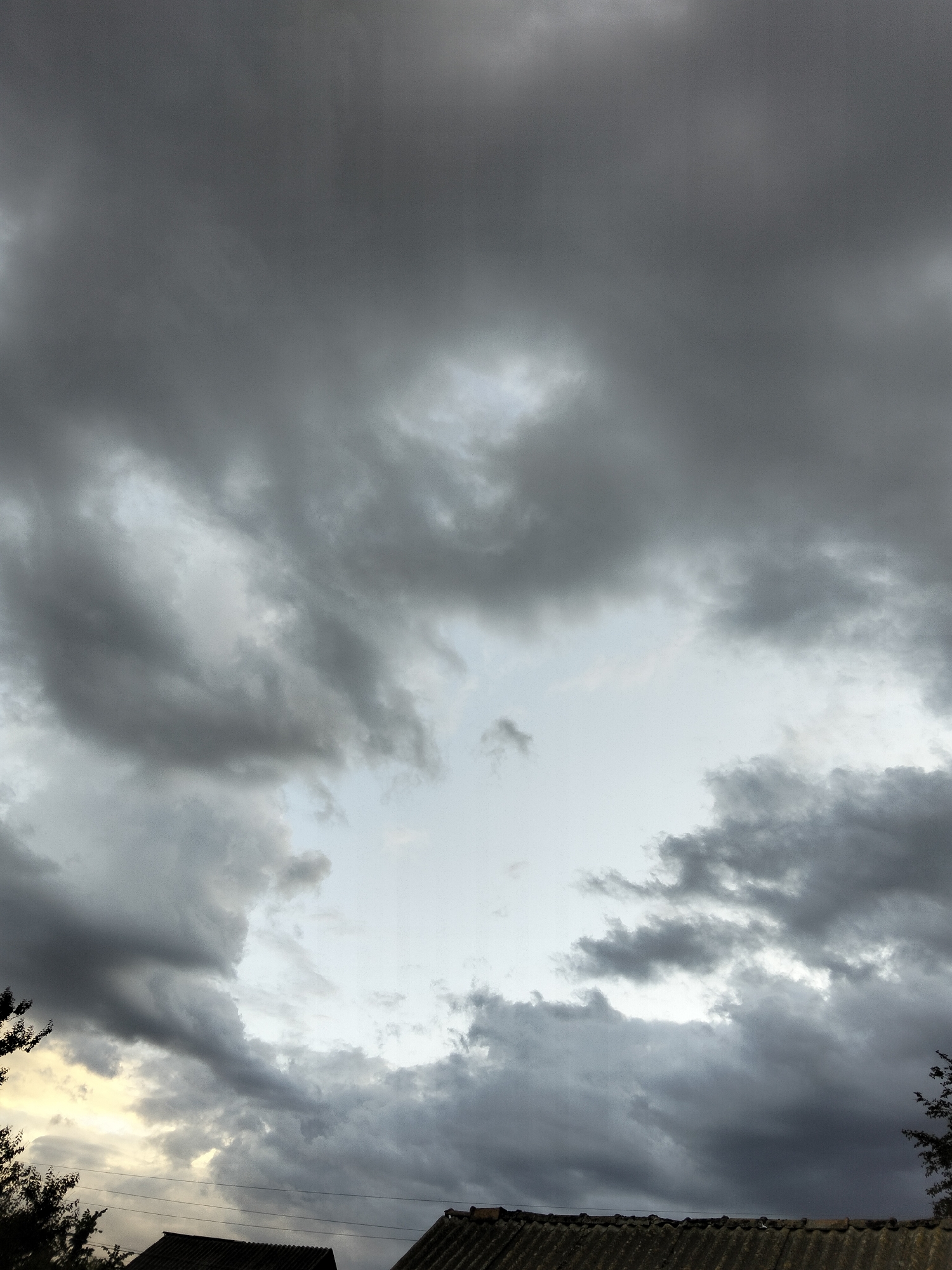The image depicts a cloudy sky over the village of Ustyvytsia, Ukraine. The sky is dominated by heavy, dark gray clouds with lighter patches in between, suggesting an overcast or approaching storm. The upper portion of the image is entirely filled with these clouds. Towards the bottom, the silhouettes of two rooftops are visible, one on the left and another on the right. The rooftop on the left appears to be made of dark corrugated material, while the one on the right is also dark and possibly tiled. Trees with dark foliage are visible along the left edge and a sliver of tree is visible on the right edge. Power lines are strung horizontally across the lower section of the image, between the trees and the rooftops. The lighting is dim, consistent with a cloudy day. There are no people or animals visible in the frame.
