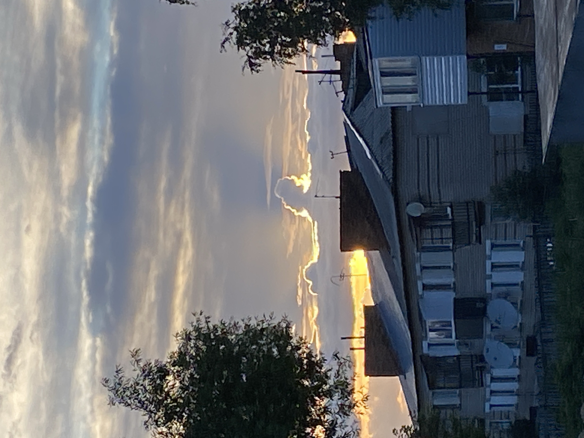 The image depicts a sunset over a residential area in Beskol Rural District, Kazakhstan. The sky is filled with dramatic clouds, with the sun breaking through a gap, casting a warm orange glow. A peculiar cloud formation, resembling a human figure with raised arms, is visible in the center of the frame, illuminated by the sunlight. In the foreground, several multi-story apartment buildings with corrugated metal roofs and satellite dishes are silhouetted against the sky. Trees flank the buildings on the left and right, their leaves catching the last rays of the sun. The scene appears to be captured from a slightly elevated viewpoint, looking across the rooftops towards the horizon.