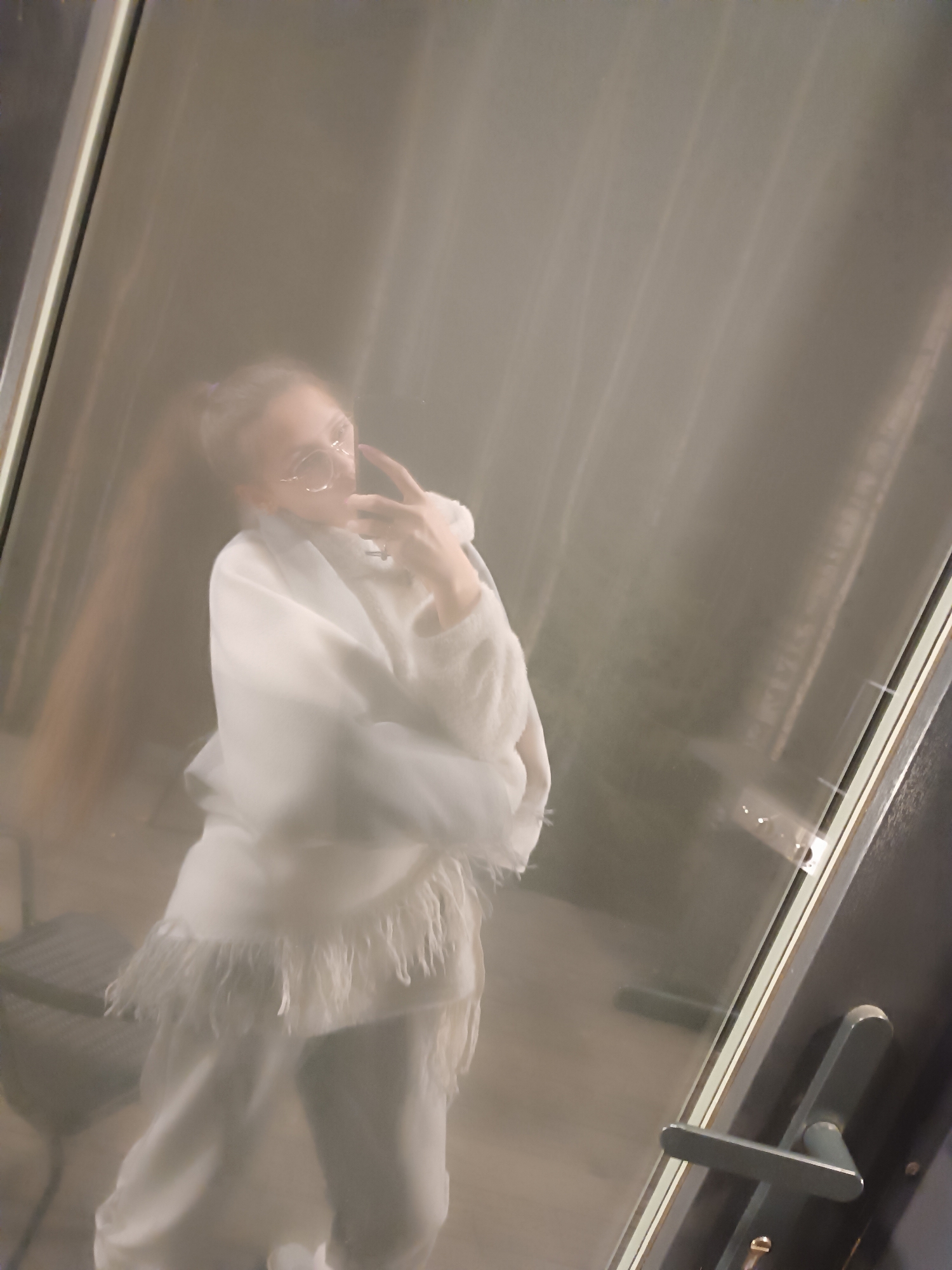 A person, possibly female, is taking a selfie in a mirror. The person is wearing glasses and a white outfit consisting of a hooded top and pants with fringe detailing. They are holding a smartphone in their right hand. The location appears to be indoors, possibly a fitting room, indicated by the mirror and door handle visible in the reflection. The exact location in Сепыч, Russia, cannot be definitively determined from the image alone.
