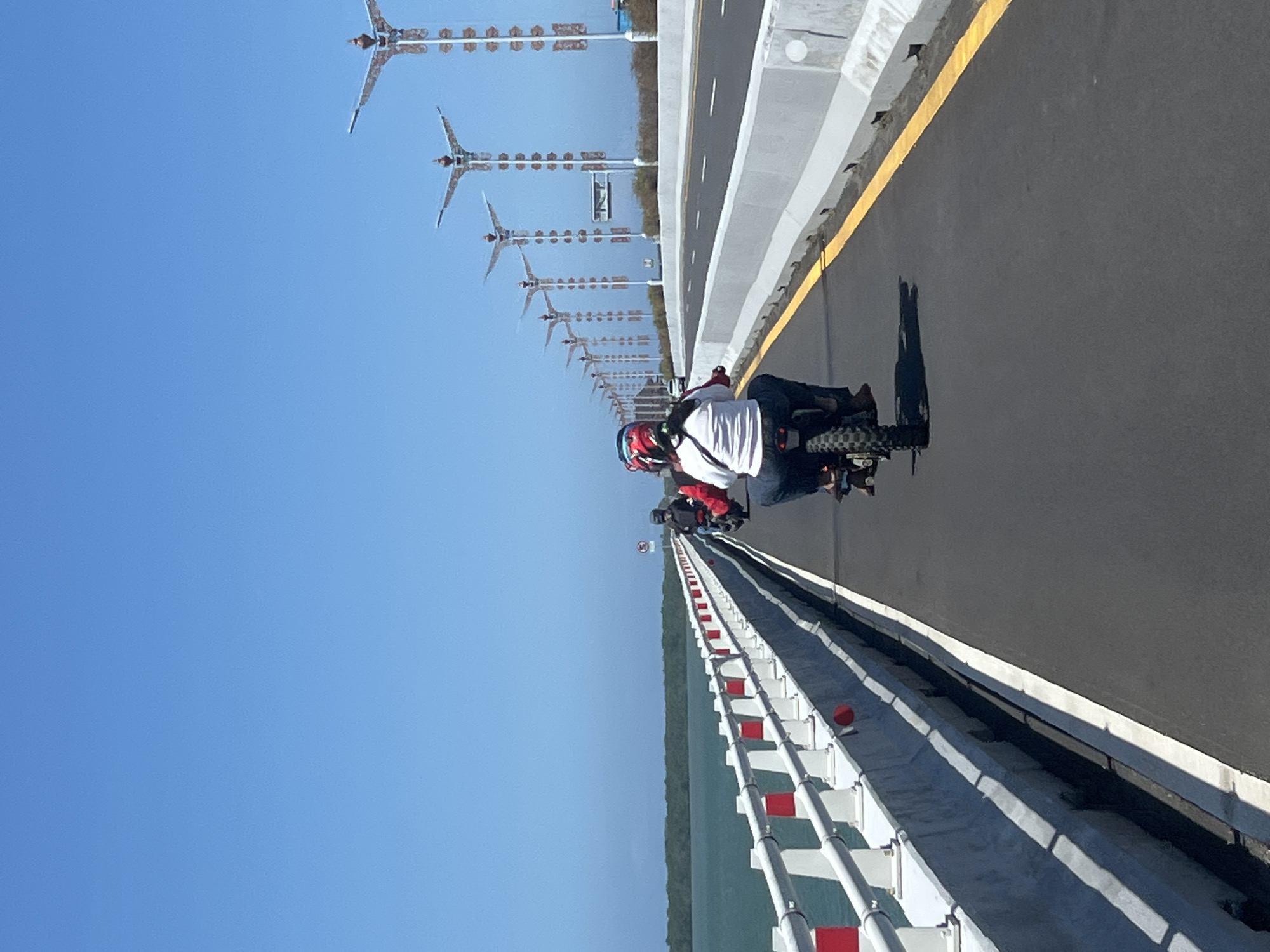 The image shows a view from the back of a motorcycle on a bridge in Kuta Selatan, Indonesia. The bridge has a railing on the left side with white posts and red and white markers. The railing separates the bridge from a body of water and a distant shoreline with lush green trees. The bridge deck is paved with asphalt and has a yellow line marking the edge of the lane. In the foreground, a person wearing a white shirt and a red and blue helmet is riding a motorcycle. Behind them, another person on a motorcycle is partially visible. The bridge has unique, decorative lampposts spaced along the right side, extending into the distance. The sky is a clear, bright blue. The overall scene depicts a sunny day on a modern bridge, likely a thoroughfare for local transportation.