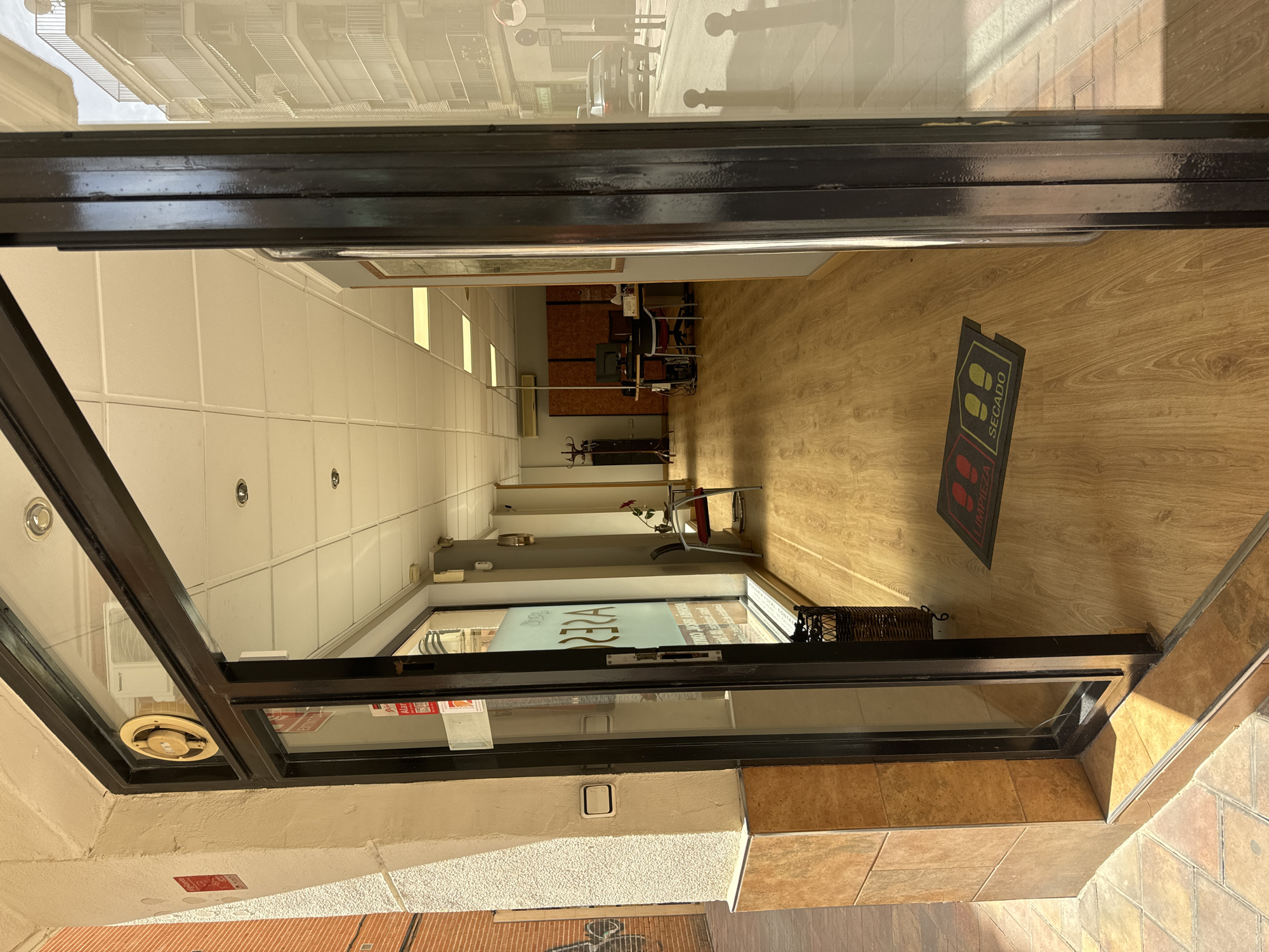 The image captures a view from outside a storefront, looking into an office or retail space. The entrance is a glass door framed by dark metal, which is partially open. The storefront appears to be located in Murcia, Spain, as indicated by the text "Limpieza" and "Secado" on a mat near the entrance, which are Spanish words for "cleaning" and "drying."
Inside, the space has a long hallway with light wood-paneled flooring. The walls are painted a neutral color, and the ceiling is tiled with recessed fluorescent lighting. On the left wall, there is a frosted glass window with a logo that appears to read "AREZA" in reverse. Further down the hallway, a desk with computer monitors and office chairs is visible. To the right, a chair with a red cushion and a small table with a flower are placed near the entrance. Sunlight streams into the space, casting shadows on the floor and creating a bright, airy atmosphere.
Outside the storefront, the sidewalk is paved with tiles, and a brick building is visible to the left. The street can be seen through the glass doors, with a car and streetlights in the distance. The overall impression is that of a clean, well-lit, and organized commercial space in an urban environment.
