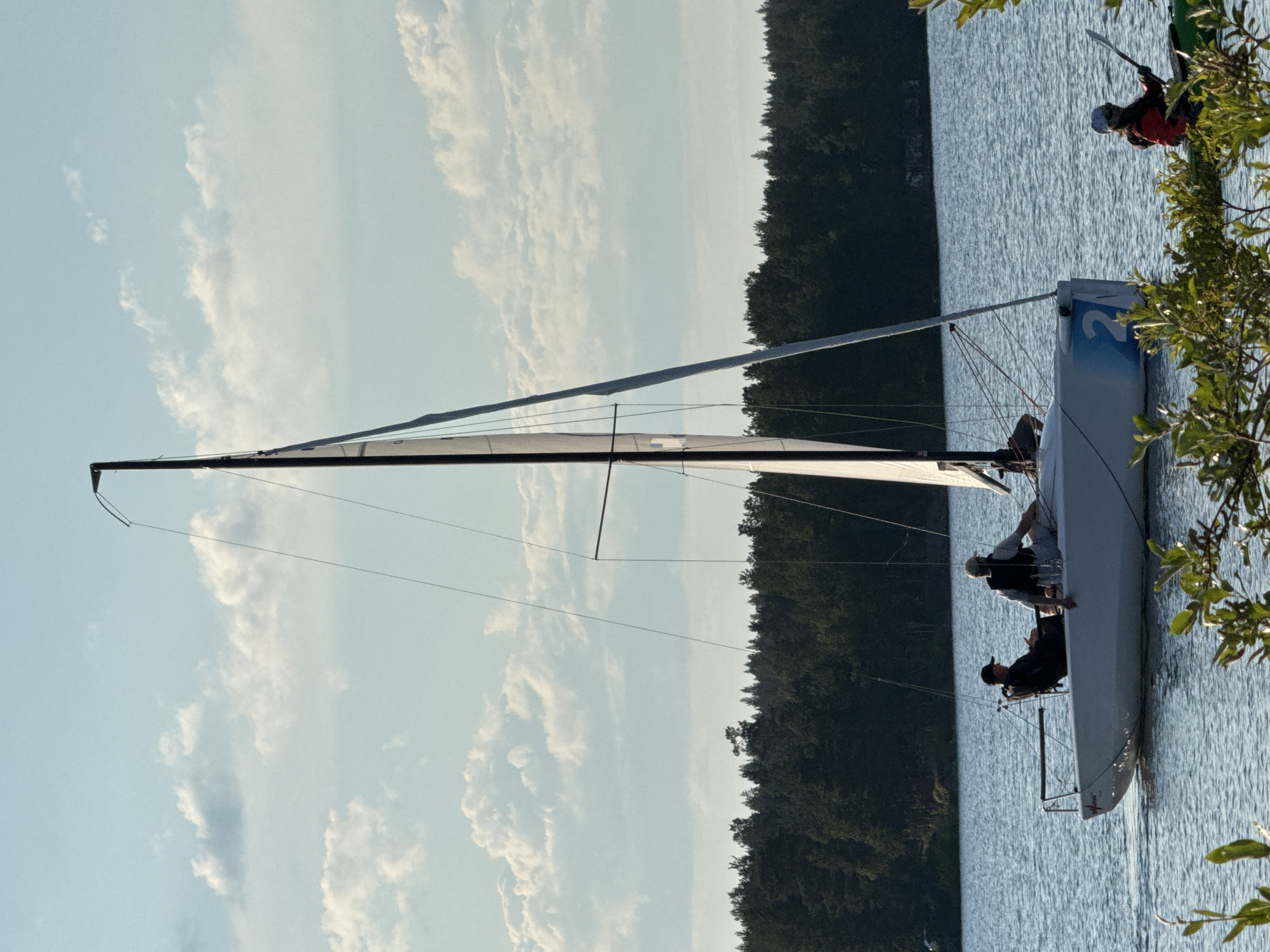 In the image, a white sailboat with a tall mast and a partial sail is on the water. Two individuals are seated on the sailboat. The person in the front is wearing a dark baseball cap, a dark shirt, and is looking towards the left. The person behind is wearing a life vest over a light-colored shirt and shorts. The sailboat has the number "2" in blue on its stern. To the right of the sailboat, a person in a green kayak with a red jacket and a helmet is paddling, holding a paddle. The kayak is in the foreground. The background shows a dark green tree line along the horizon, with a light blue sky filled with scattered white clouds above. The water appears calm with ripples. The scene is outdoors, likely on a lake or calm body of water.