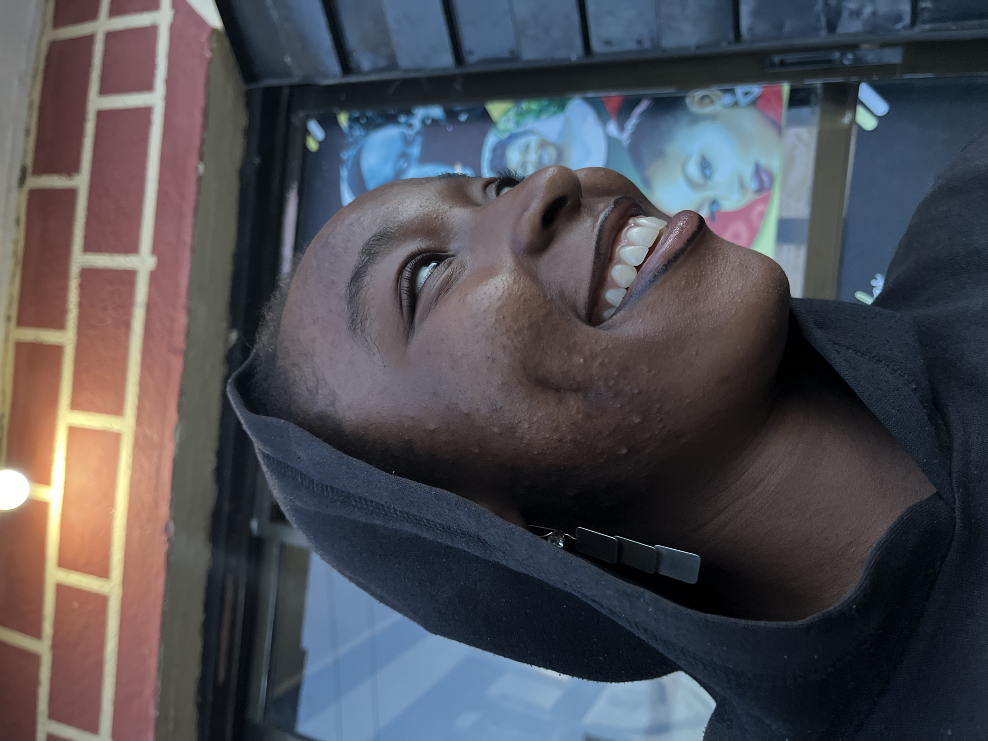 A young individual with dark skin, wearing a black hooded sweatshirt, is depicted in a close-up, chest-up shot. The person is positioned facing right in the frame, looking upwards and to the right with a wide, genuine smile, revealing white teeth and prominent dimples. The left eye is partially visible, crinkled with the smile. A long, dark, rectangular dangle earring with a small light-colored top element is visible on their left ear, extending from beneath the hood. The skin on their face and neck shows natural texture, including visible pores and some minor bumps.
The background consists of a reddish-brown brick wall with lighter horizontal mortar lines or painted stripes at the top of the frame. A single, bright, circular light fixture is mounted on this wall near the top center. Below the brickwork, a dark, horizontal structural element is visible. To the right of the subject, a dark, slatted or paneled door or shutter is partially visible. Behind the subject, a reflective surface, likely a glass window or door, displays blurred, colorful images. These appear to be posters or advertisements featuring stylized portraits, predominantly women, with distinct blue, red, and green color schemes. One prominent blue-toned face is visible at mid-level, with another blurred face above it and a smiling face lower down. The overall setting appears to be an outdoor urban or commercial environment.