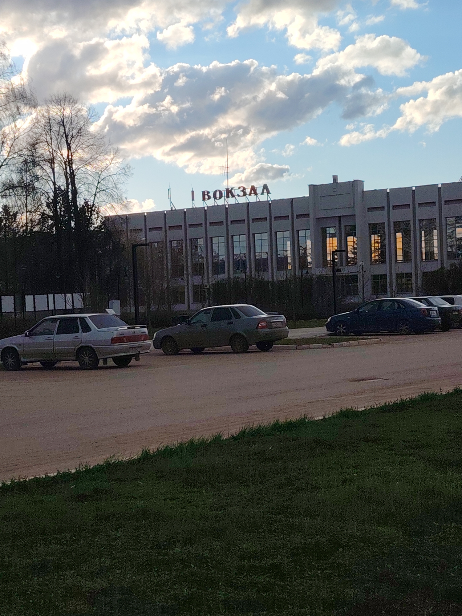 The image presents an outdoor scene in Yanaul, Russia, featuring a large, multi-story building, likely a transport station, as indicated by the prominent red Cyrillic text "ВОКЗАЛ" (VOKZAL, meaning "Station") displayed on its roofline. The building exhibits a robust architectural style, characterized by a grey facade with vertical pillar-like structures and numerous rectangular windows. Reflections of warm, possibly setting or rising sun, cast an orange glow on some of the windows.
In front of the building, several cars are parked on what appears to be a dirt or gravel lot. In the foreground, a light-colored sedan (resembling a Lada model) and a darker, possibly olive-green sedan, are clearly visible. Additional cars are parked further along the lot. To the left of the building and behind the parked cars, there is dense foliage and trees, many appearing bare or sparsely leafed, suggesting either early spring or late autumn. A strip of green grass occupies the bottom portion of the foreground.
The sky above is a bright blue with numerous large, puffy white and grey cumulus clouds, indicating fair weather conditions. The overall lighting suggests either the golden hour of late afternoon or early morning. The scene appears calm and devoid of people, capturing a quiet moment at what is presumably a public transport hub.
