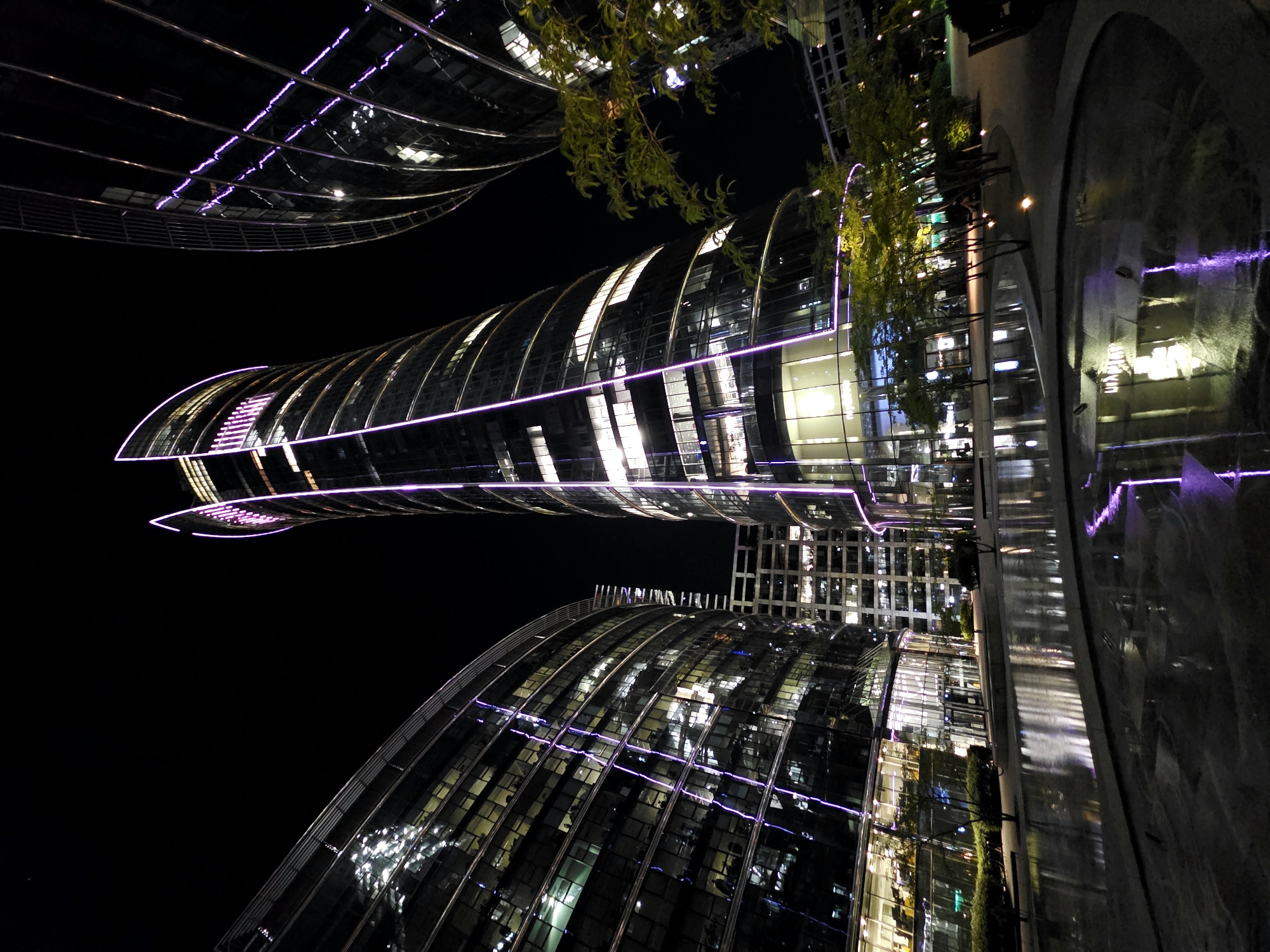 The image shows a nighttime view of the Warsaw Spire, a modern skyscraper located in the city of Warsaw, Poland. The skyscraper is illuminated with purple LED lights along its edges, creating a striking visual effect against the dark night sky. The building's glass facade reflects the lights and the surrounding cityscape. In the foreground, there is a reflective pool of water, possibly a fountain, which mirrors the illuminated buildings. Several trees with green foliage are also visible, adding a touch of nature to the urban landscape. The overall scene depicts a vibrant and contemporary urban environment.