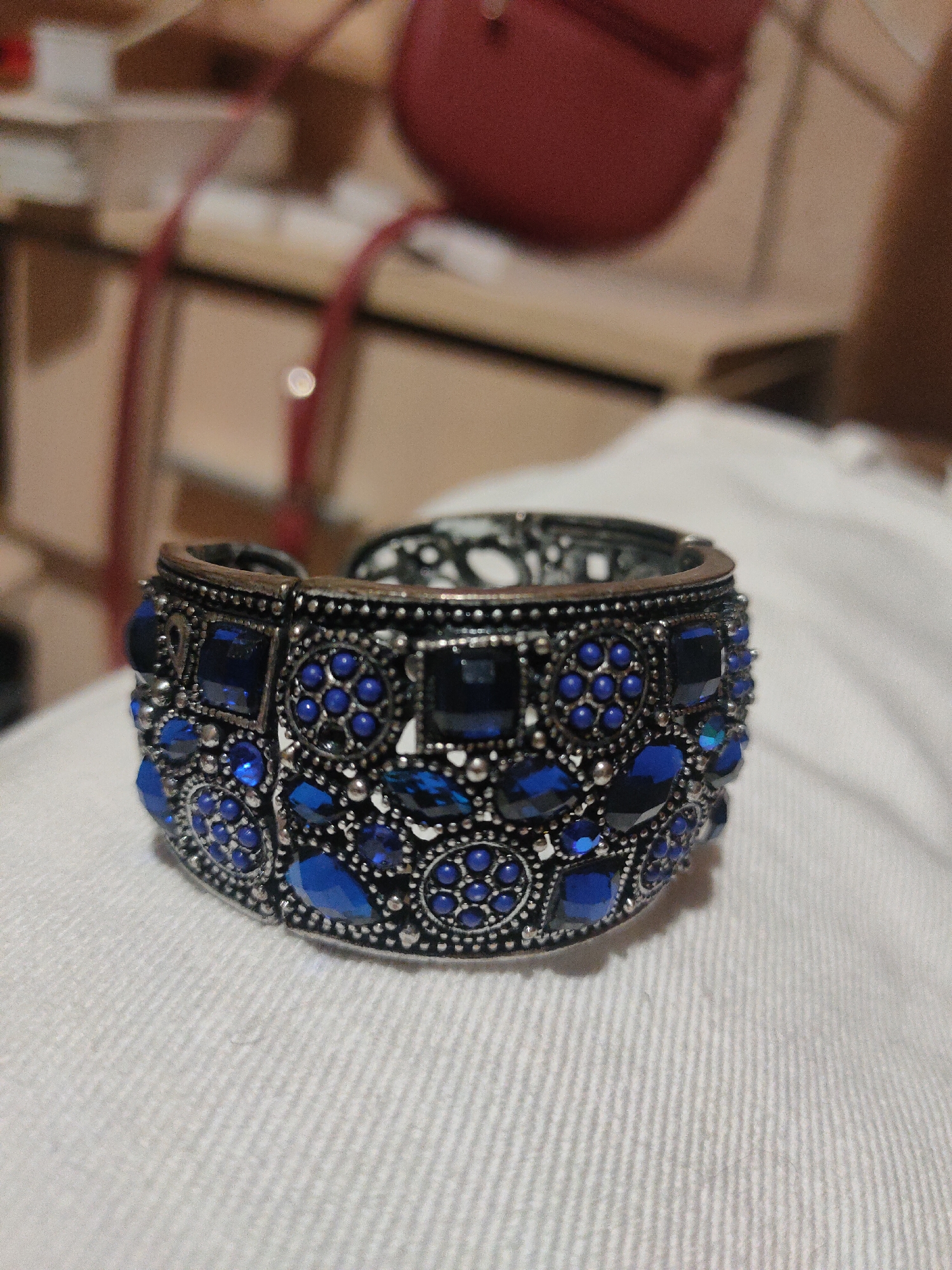 A close-up, eye-level shot focuses on a wide, ornate silver cuff bracelet adorned with various shapes and sizes of faceted blue gemstones. The bracelet is resting on a textured white surface, likely fabric. The background is blurred and indistinct, showing hints of furniture and a red bag, suggesting an indoor setting. The image is sharp and well-lit, highlighting the intricate details of the bracelet.