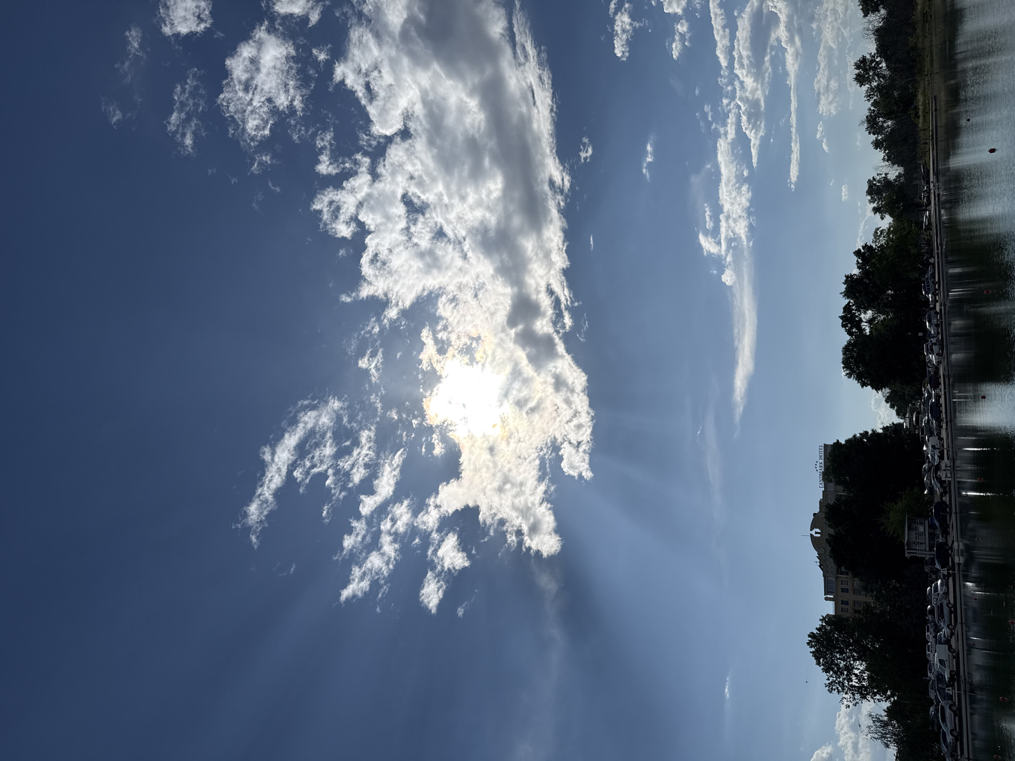 The image depicts a partially cloudy sky above the Landmark Hotel Plovdiv in Bulgaria. The sun is partially obscured by white clouds, creating sunbeams through the sky. The hotel is visible with trees in front, next to a body of water reflecting the scene. A row of parked cars is present next to the water, adjacent to the trees.