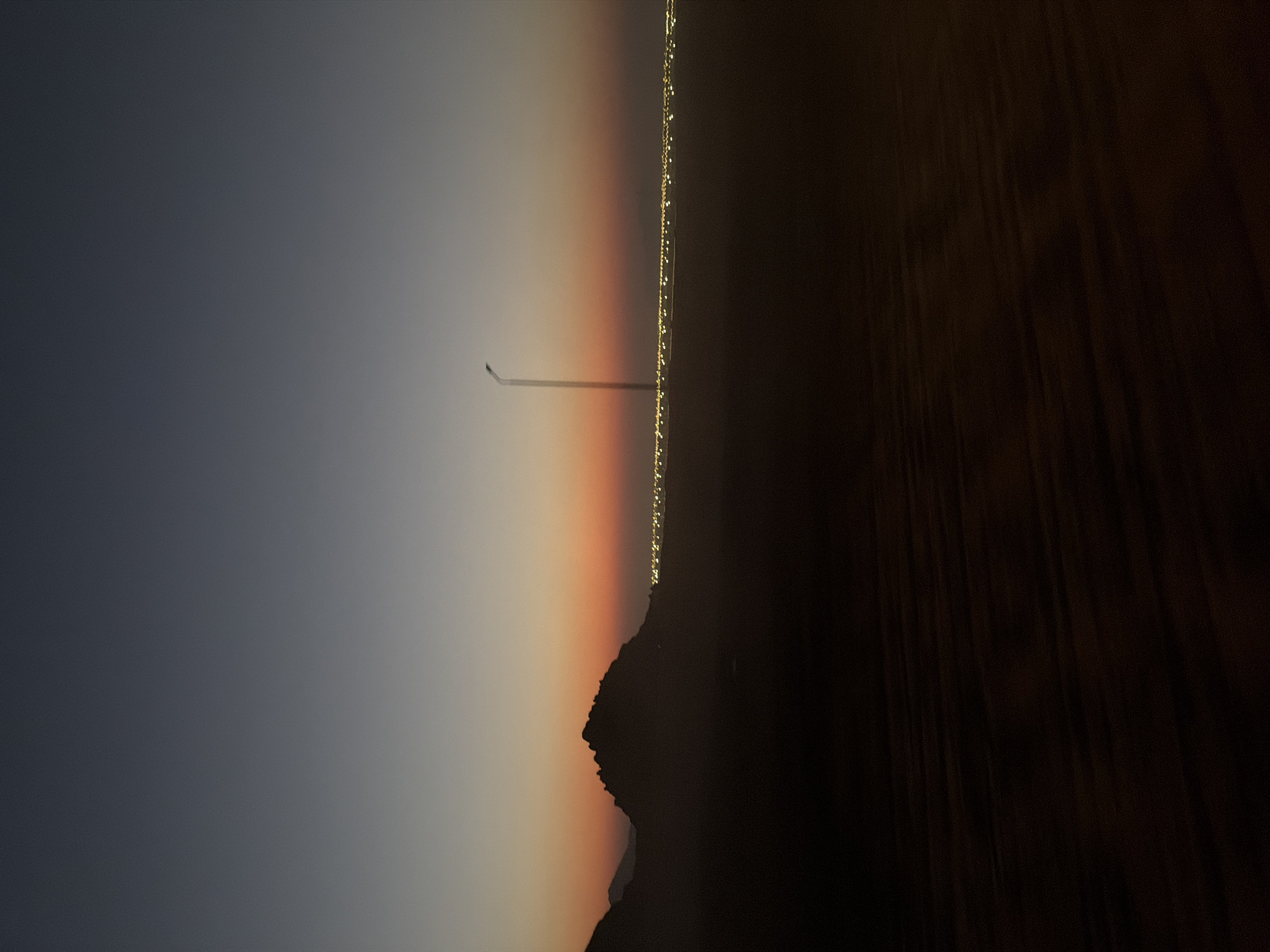 The image depicts a dimly lit desert landscape with a distant city illuminated by lights. The sky transitions from a dark blue at the top to a gradient of orange and yellow near the horizon, suggesting either dawn or dusk. In the foreground, dark, undulating sand dunes create a stark contrast with the brighter background. To the left, a silhouette of a rugged mountain range is visible against the colorful sky. A lone street lamp stands tall on the right side, its light barely visible against the ambient light. The city lights stretch horizontally across the middle ground, appearing as a faint, glowing line. The overall scene conveys a sense of vastness and tranquility, with the city lights offering a hint of human presence in the otherwise barren environment. It is not possible to determine the specific location within Sharm El Sheikh, Egypt, from the provided image.