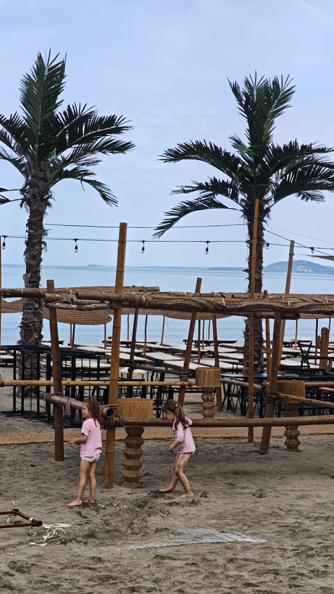 The image shows a beach scene, potentially in Burgas, Bulgaria, based on the coastal setting and architectural style. Two young girls, barefoot and wearing light pink shirts and white shorts, are the central subjects. They appear to be playing on the sand. In the background, there's a constructed outdoor structure, possibly for a beach bar or restaurant, built with bamboo-like posts and thatched roofing. Tables and chairs are visible under the structure. Two palm trees frame the scene. Overhead, string lights are hung between the palm trees and parts of the structure. In the distance, there's a body of water, presumably the Black Sea, and a faint outline of land on the horizon. The overall impression is a relaxed, seaside atmosphere.